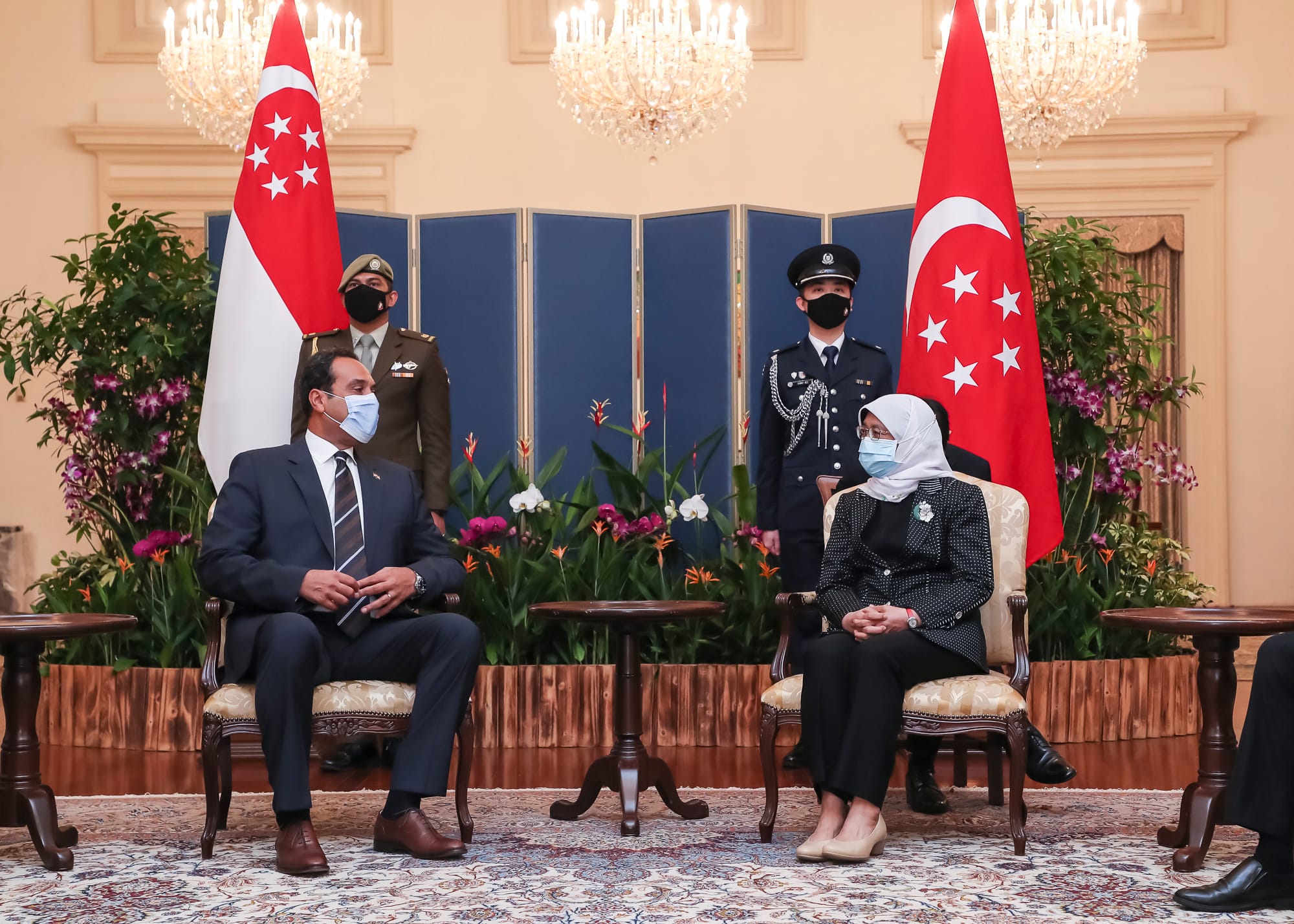 Dignitaries in formal attire seated by Singapore flags, guards stand behind.