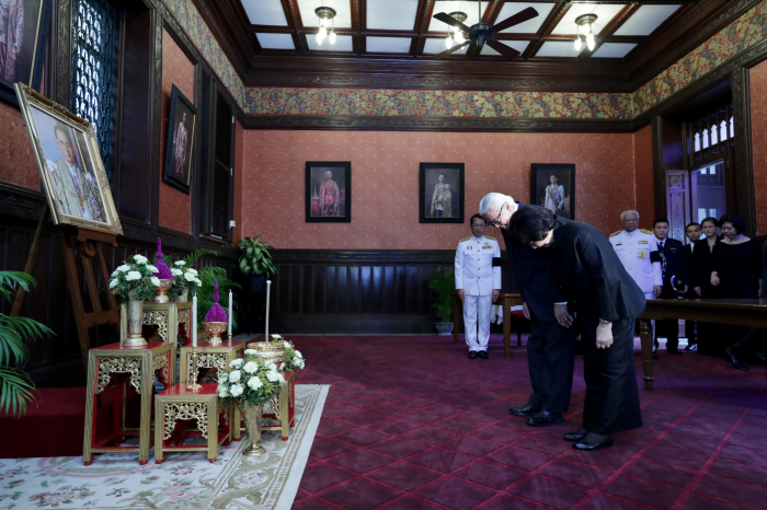 Two people bow before a framed portrait amidst floral displays in an ornate room.