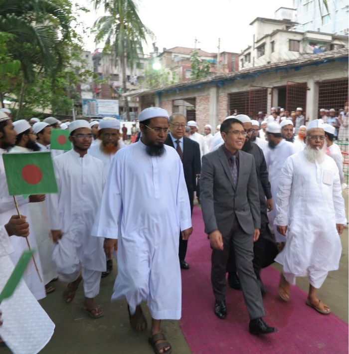 Men in white robes and caps walk with men in suits; some hold Bangladesh flags.