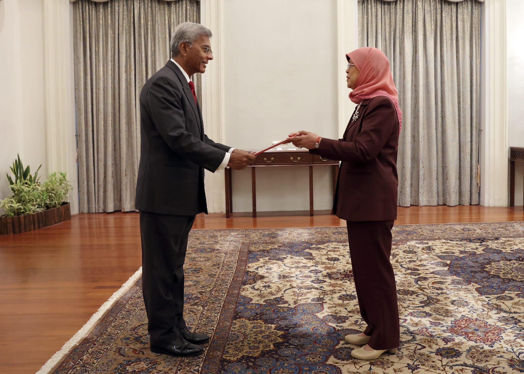 Halimah Yacob and man in suits exchanging red document in ornate room.