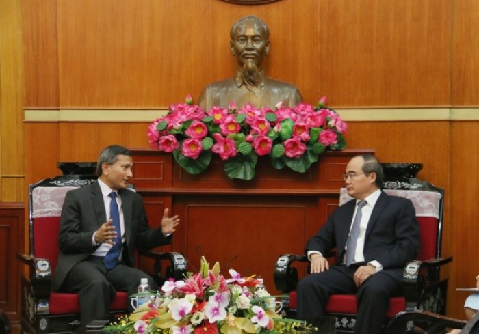 Two men in suits sit facing each other below a Ho Chi Minh bust adorned with pink lotus flowers.