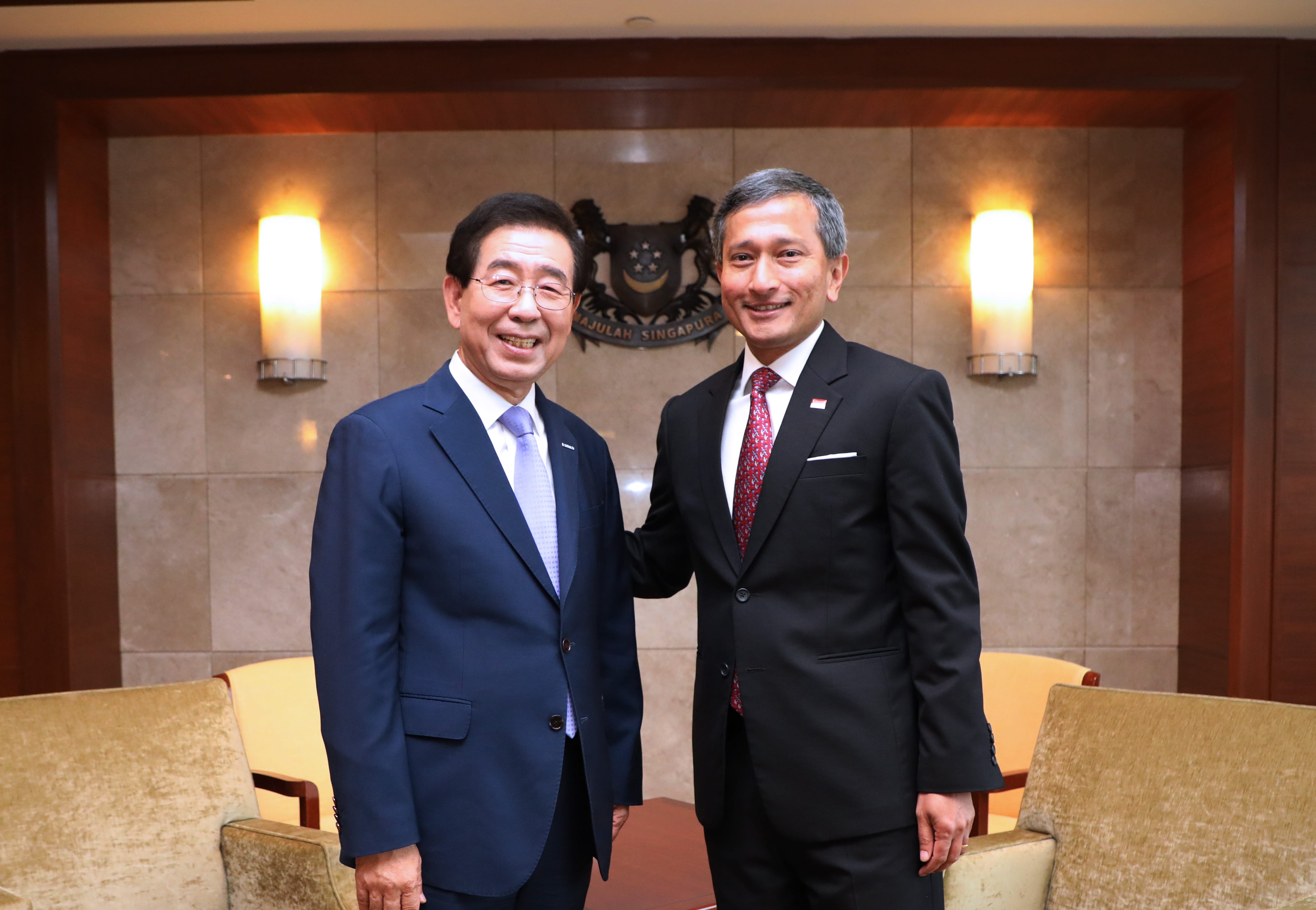 Two men in suits pose in front of a Singapore coat of arms.