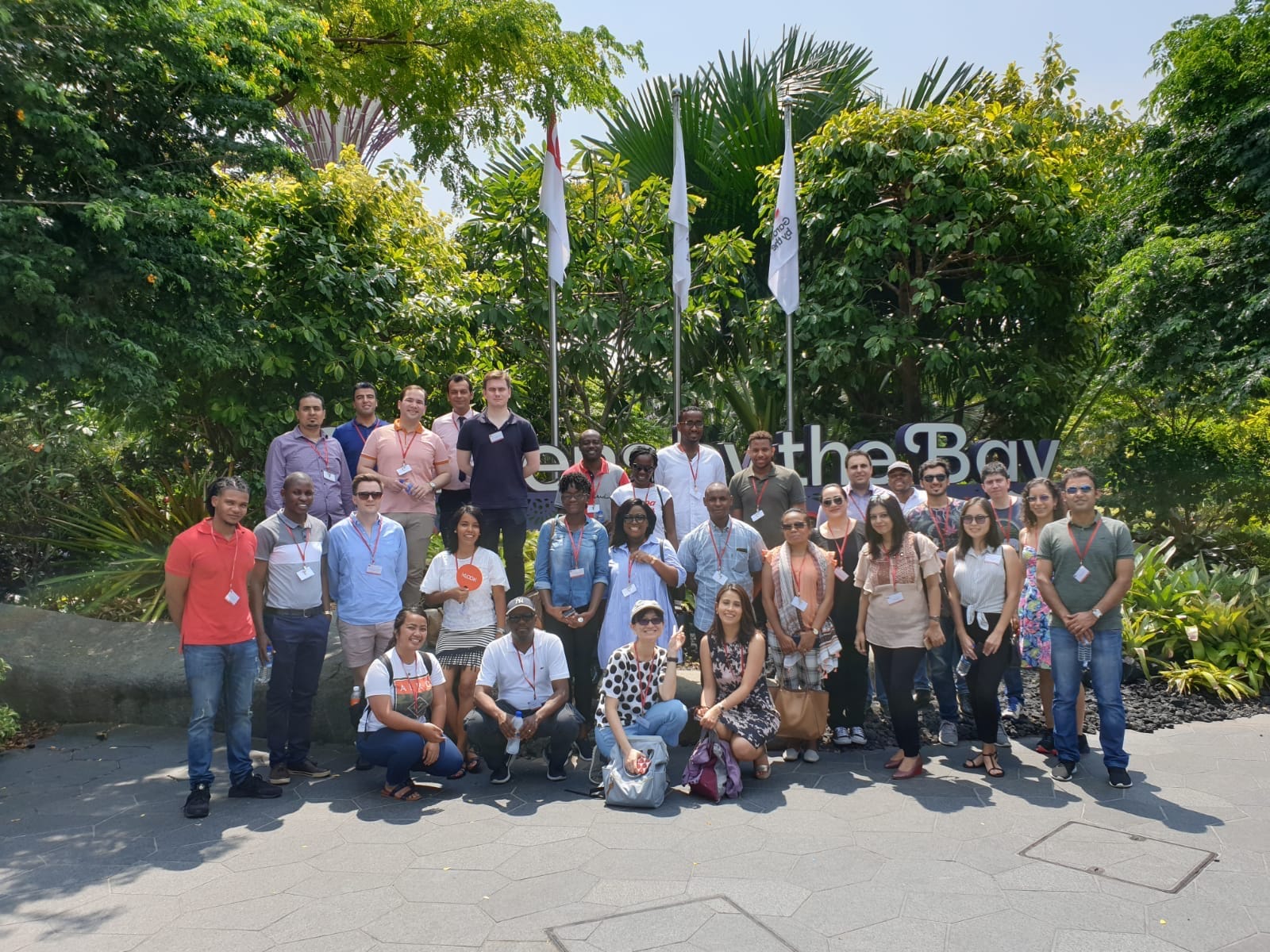 Group of about 30 people posing outdoors by "Gardens by the Bay" signage and flagpoles.
