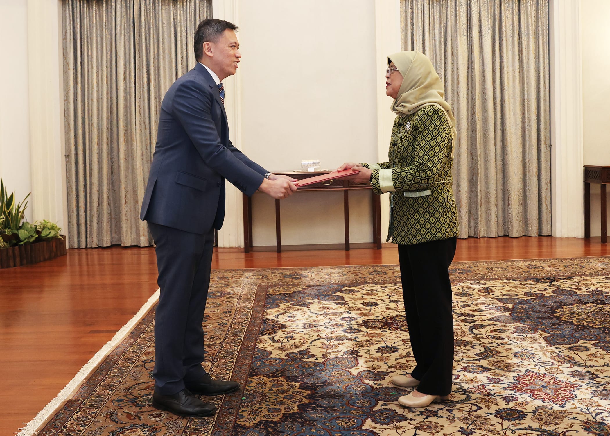 Man in suit hands file to Halimah Yacob, President of Singapore, in a room with a patterned rug.