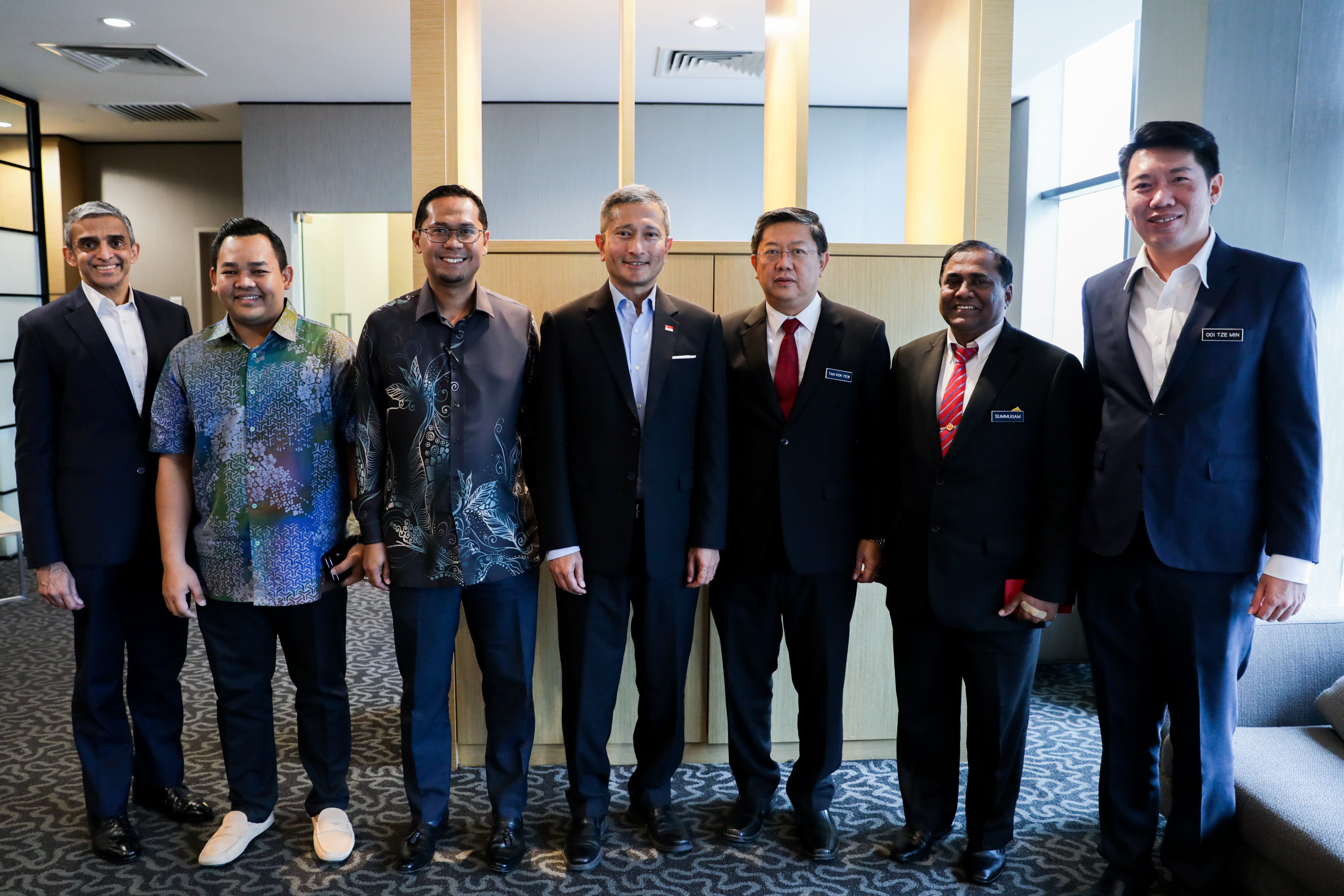 Seven men in suits or patterned shirts stand side-by-side, some wearing name tags.
