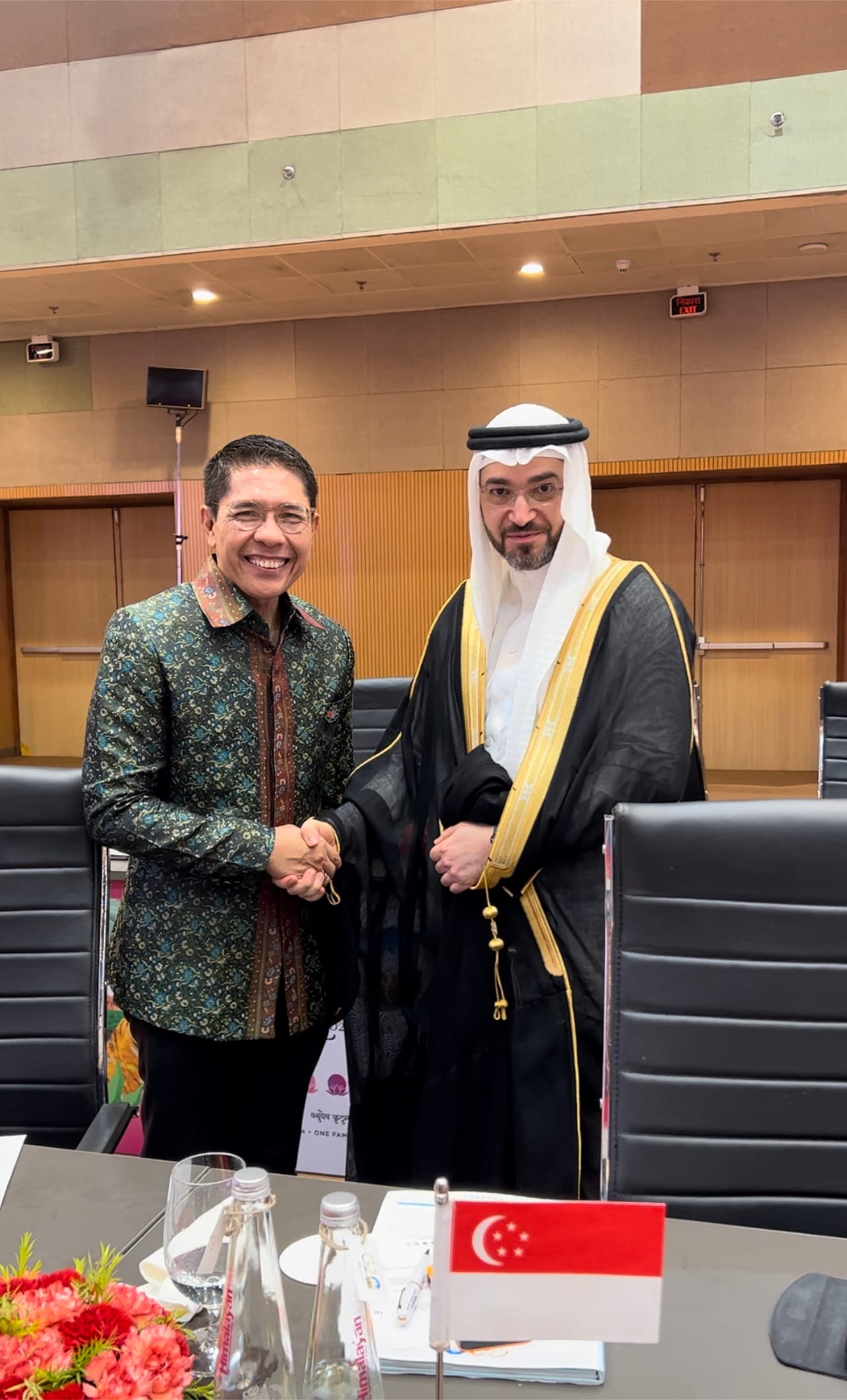 Two men in formal attire shaking hands, Singapore flag on table.
