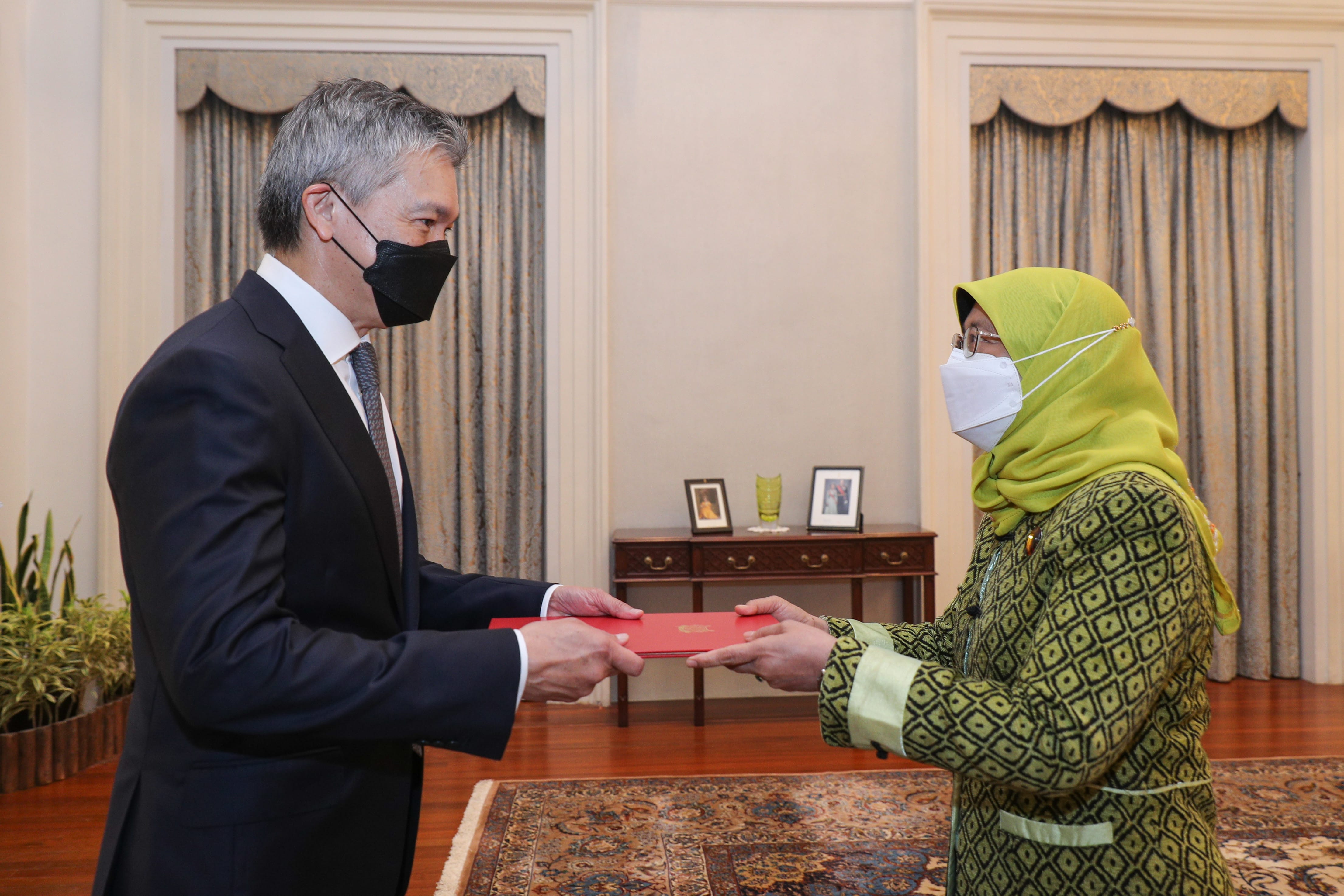 Man in suit and mask presents red folder to woman wearing hijab, glasses, and mask.