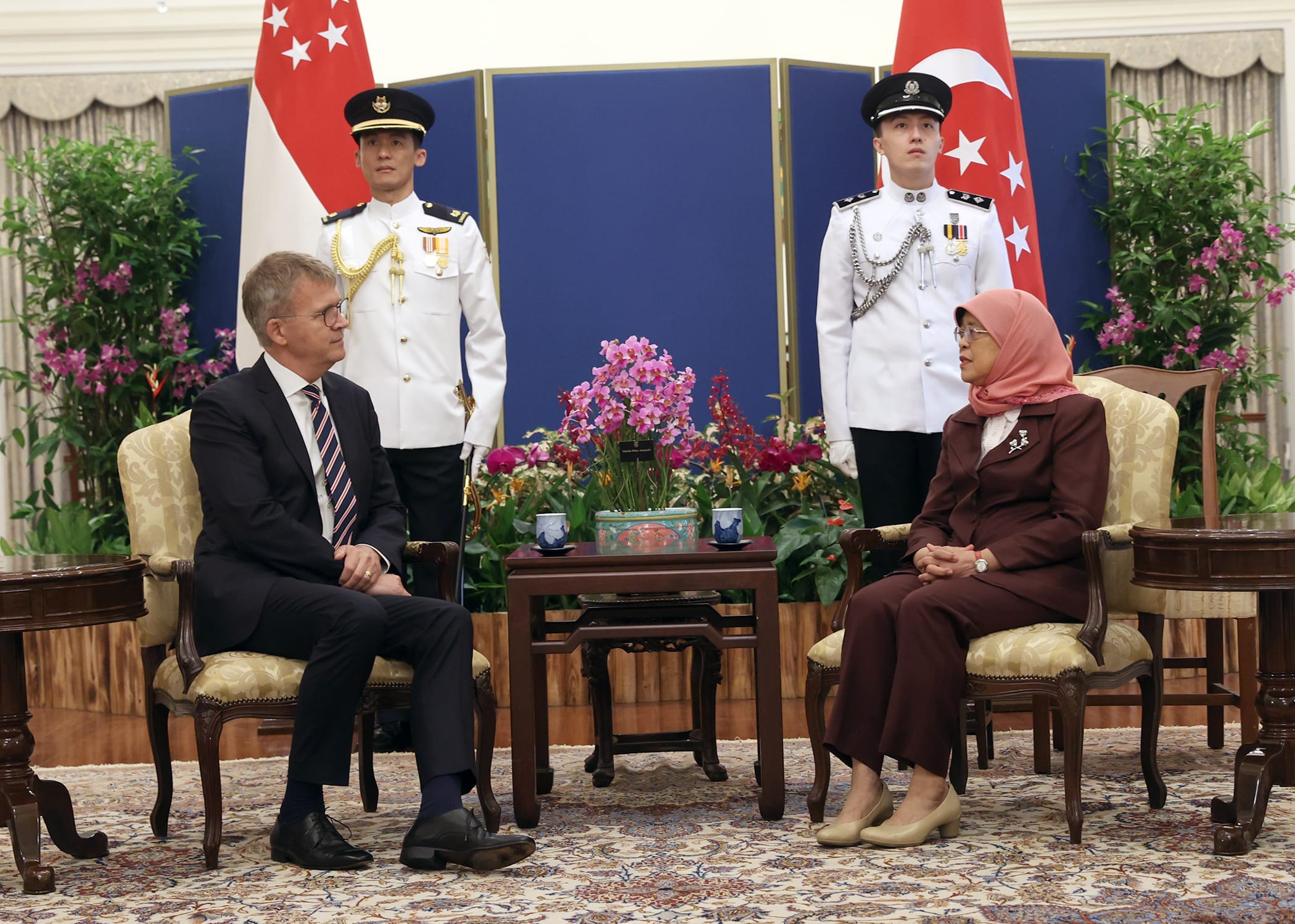 Man and woman seated, flanked by uniformed officers and Singapore flags.