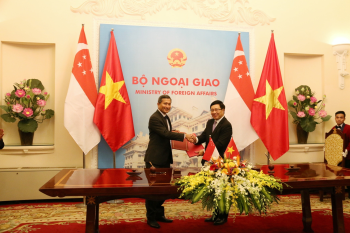 Two men shake hands between Singapore and Vietnam flags, over a table with floral arrangements.