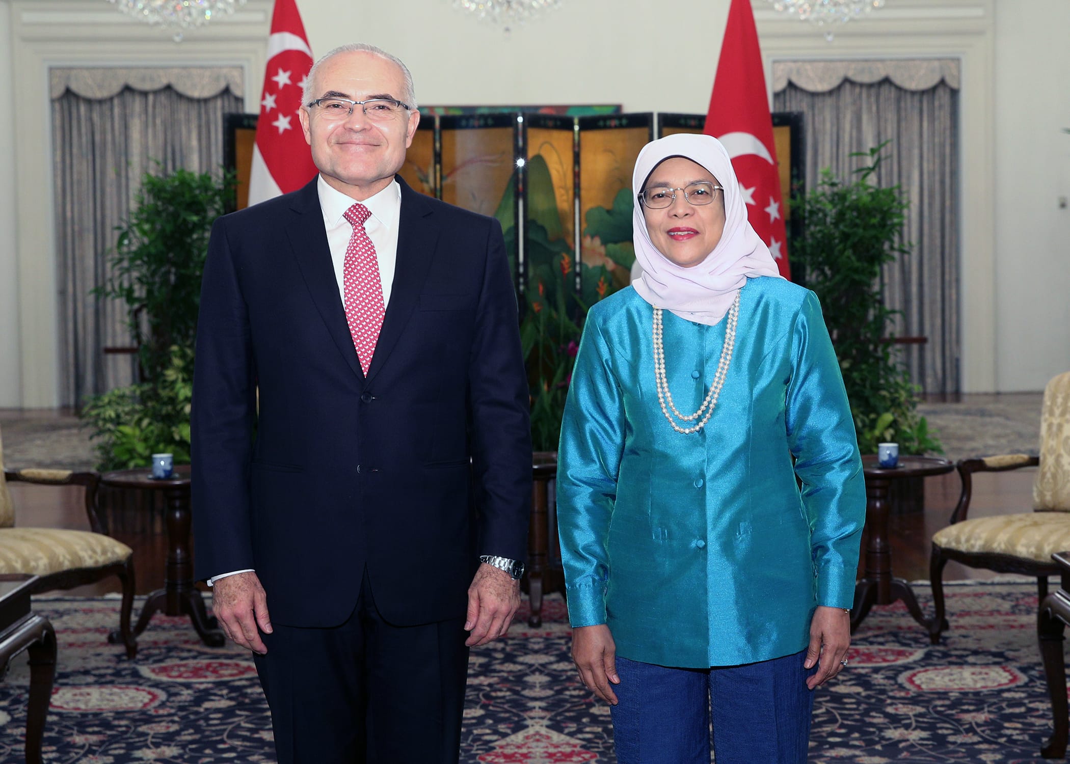 Two people standing indoors with Singapore flags behind them. Man in suit and woman in headscarf and turquoise top.