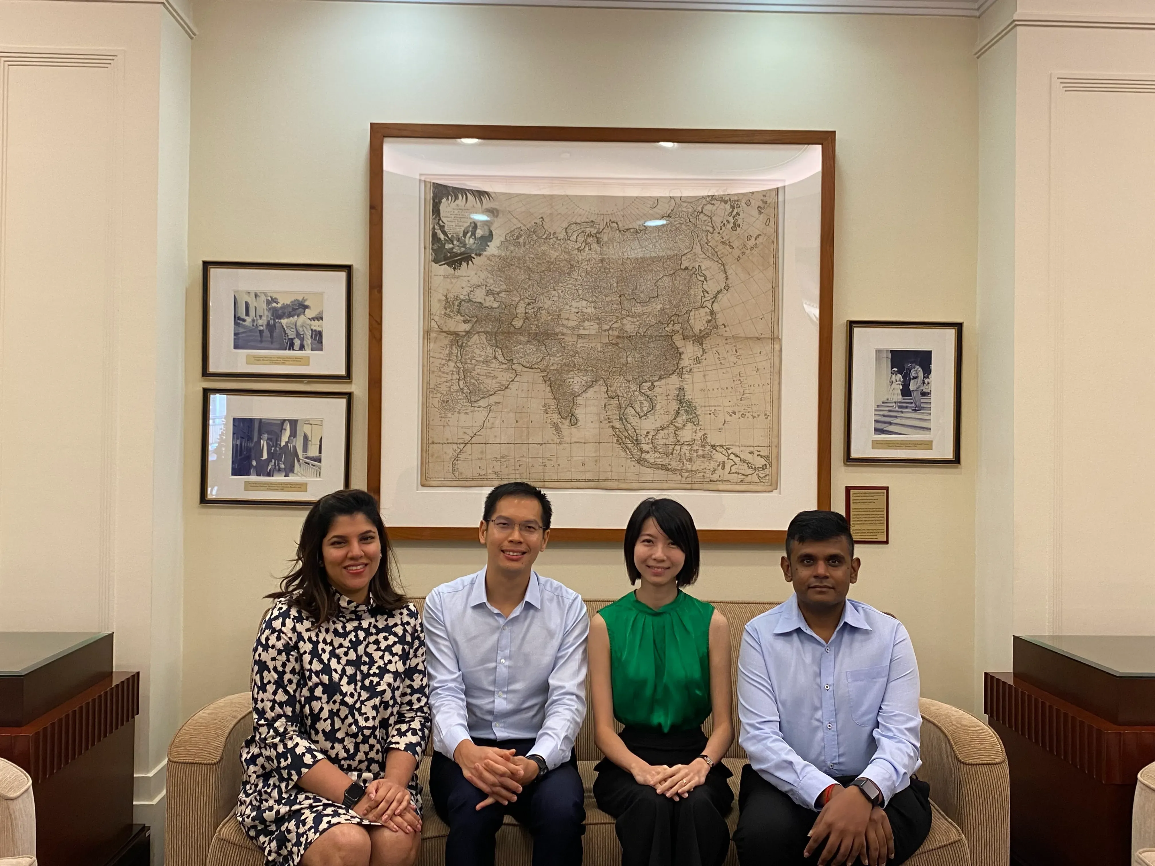A group of four people seated on a couch in front of a framed map and photos on the wall.