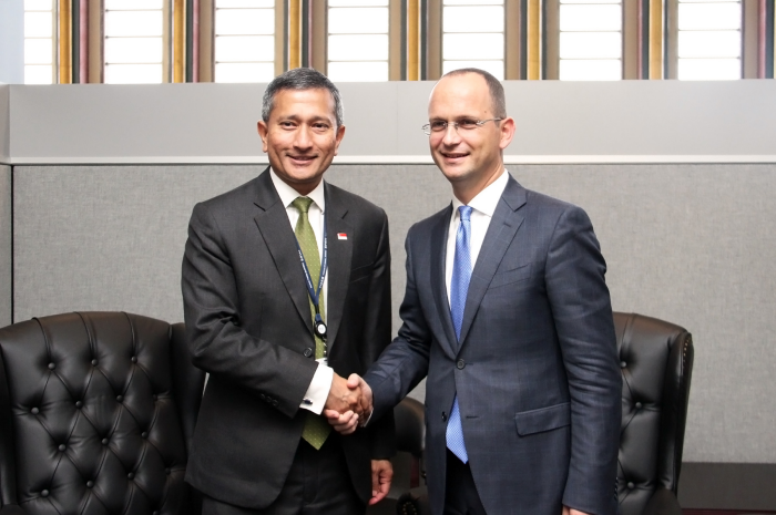 Two men in suits shaking hands in front of a gray wall and tufted black chairs.