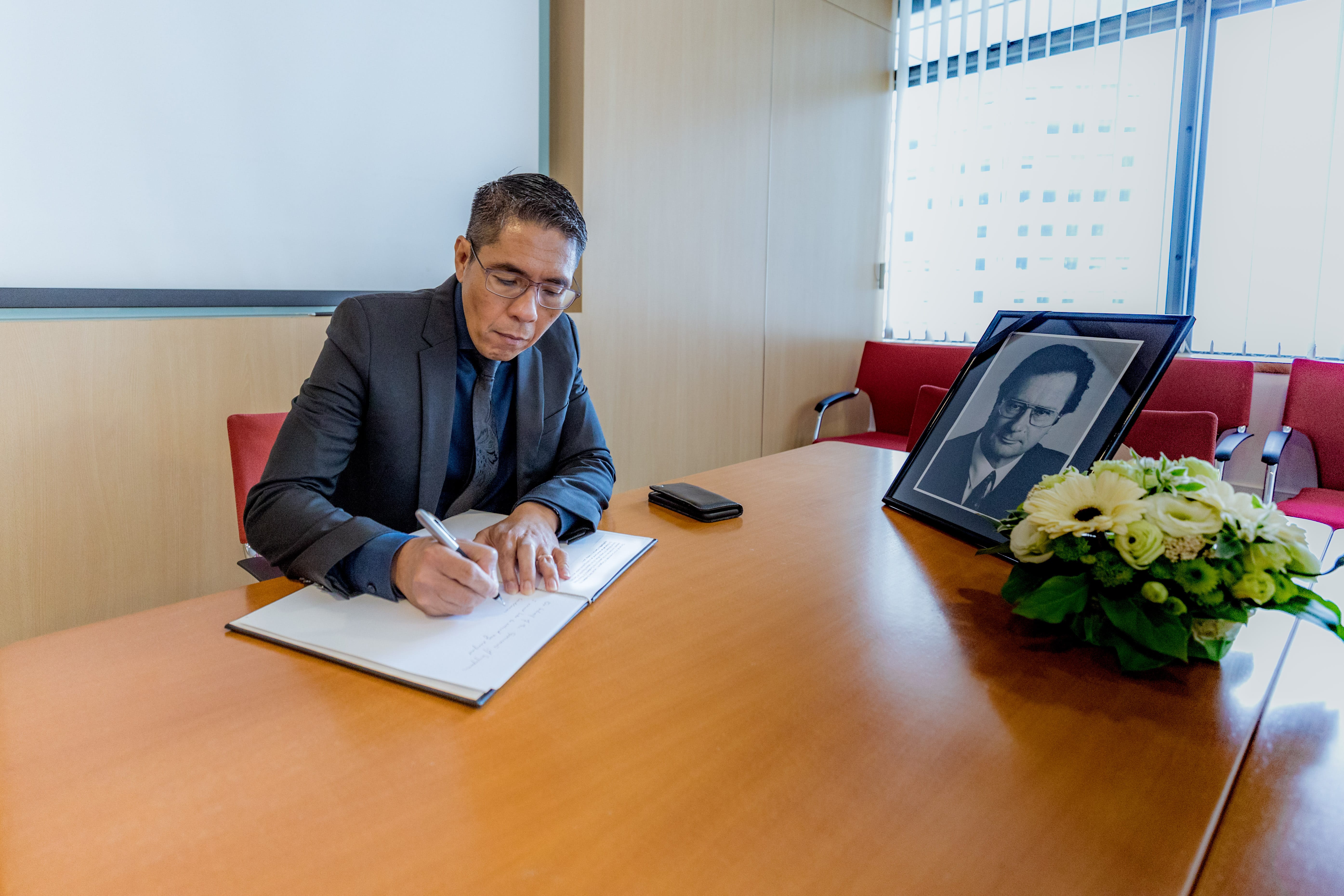 Man in suit signing book at a table with a framed photo and flowers.