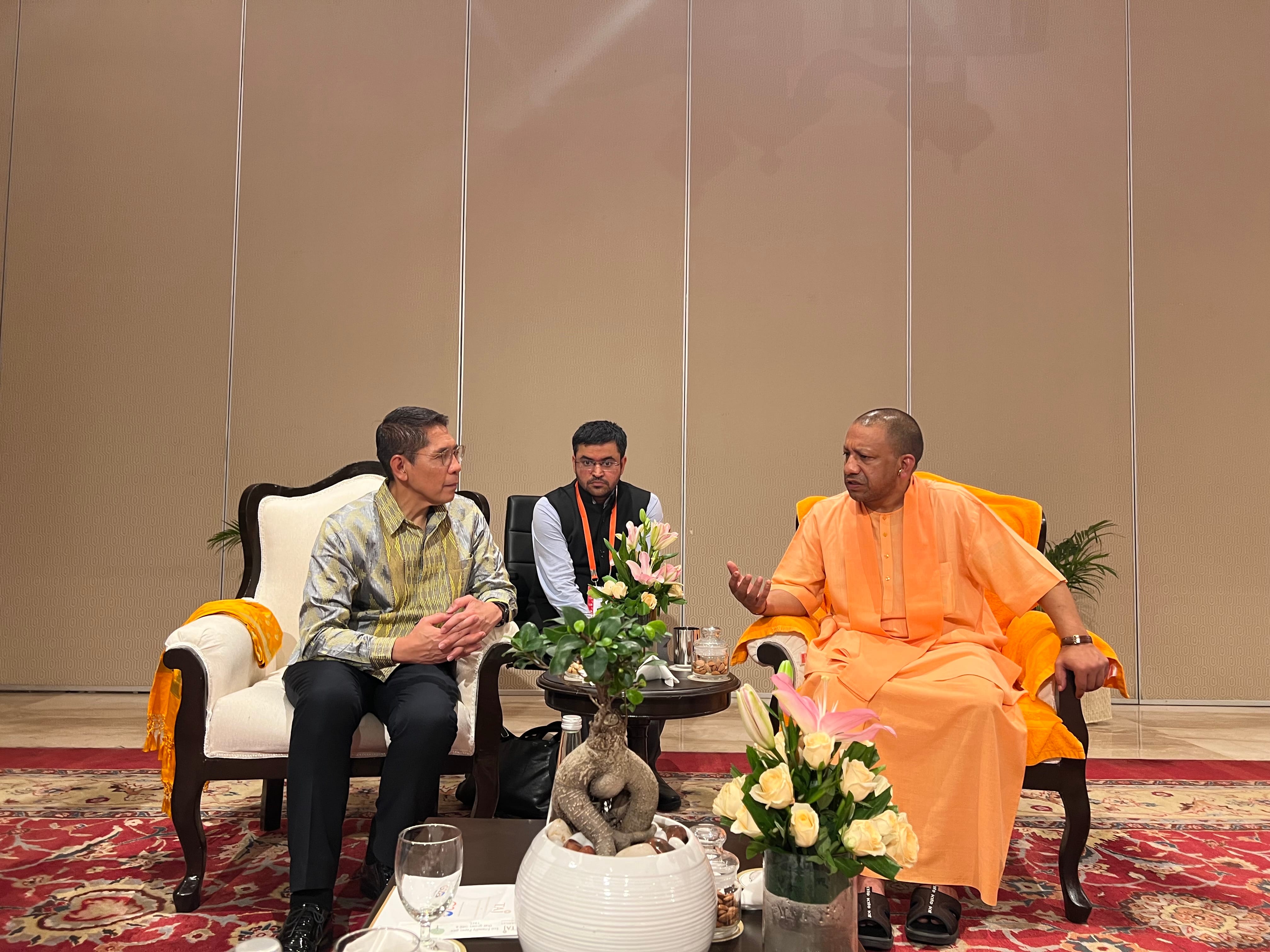 Three men seated in chairs face-to-face with a floral centerpiece on a table. One man is wearing orange robes.