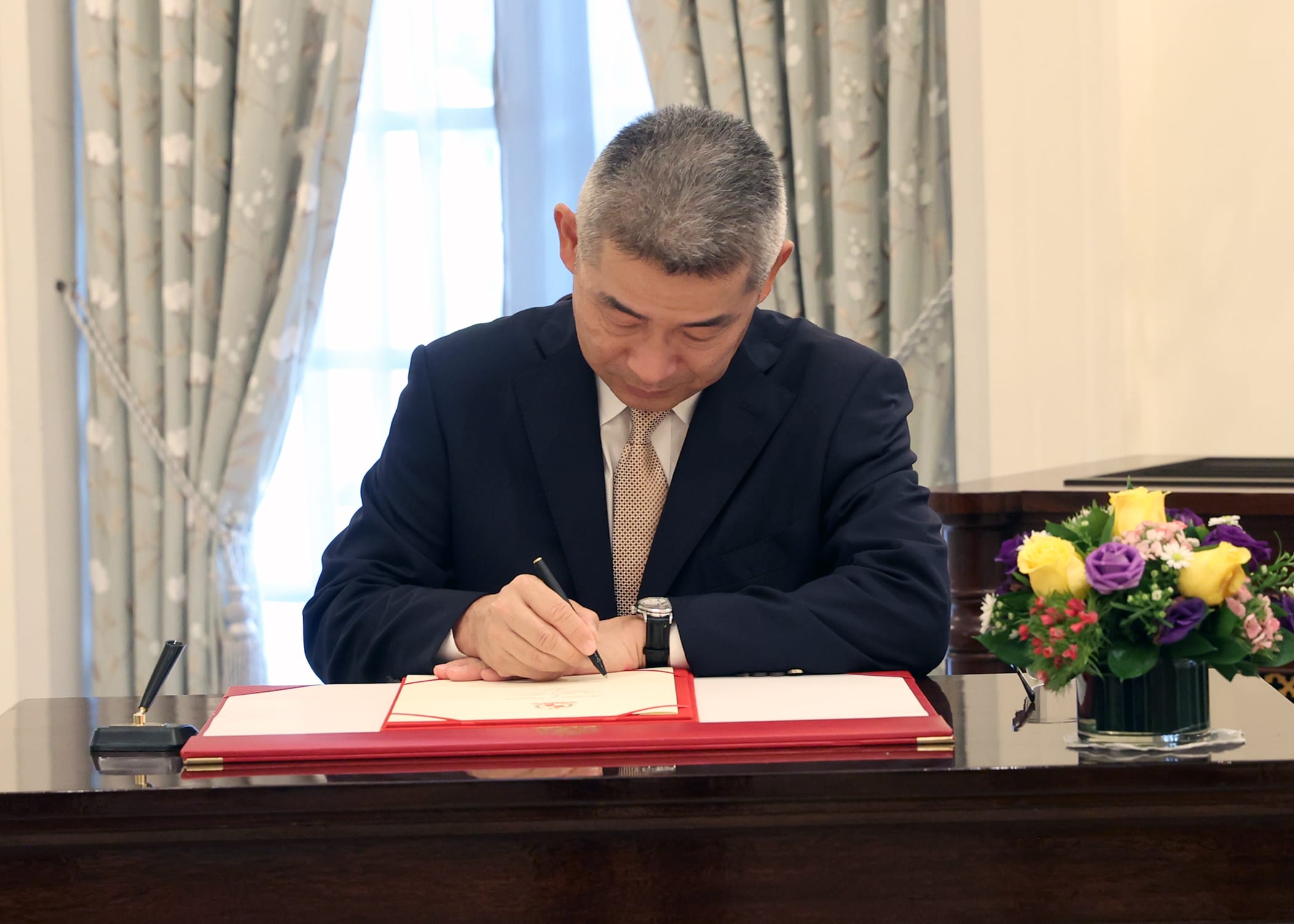 Man in suit signing document with pen. Flowers on desk. Draped window behind.