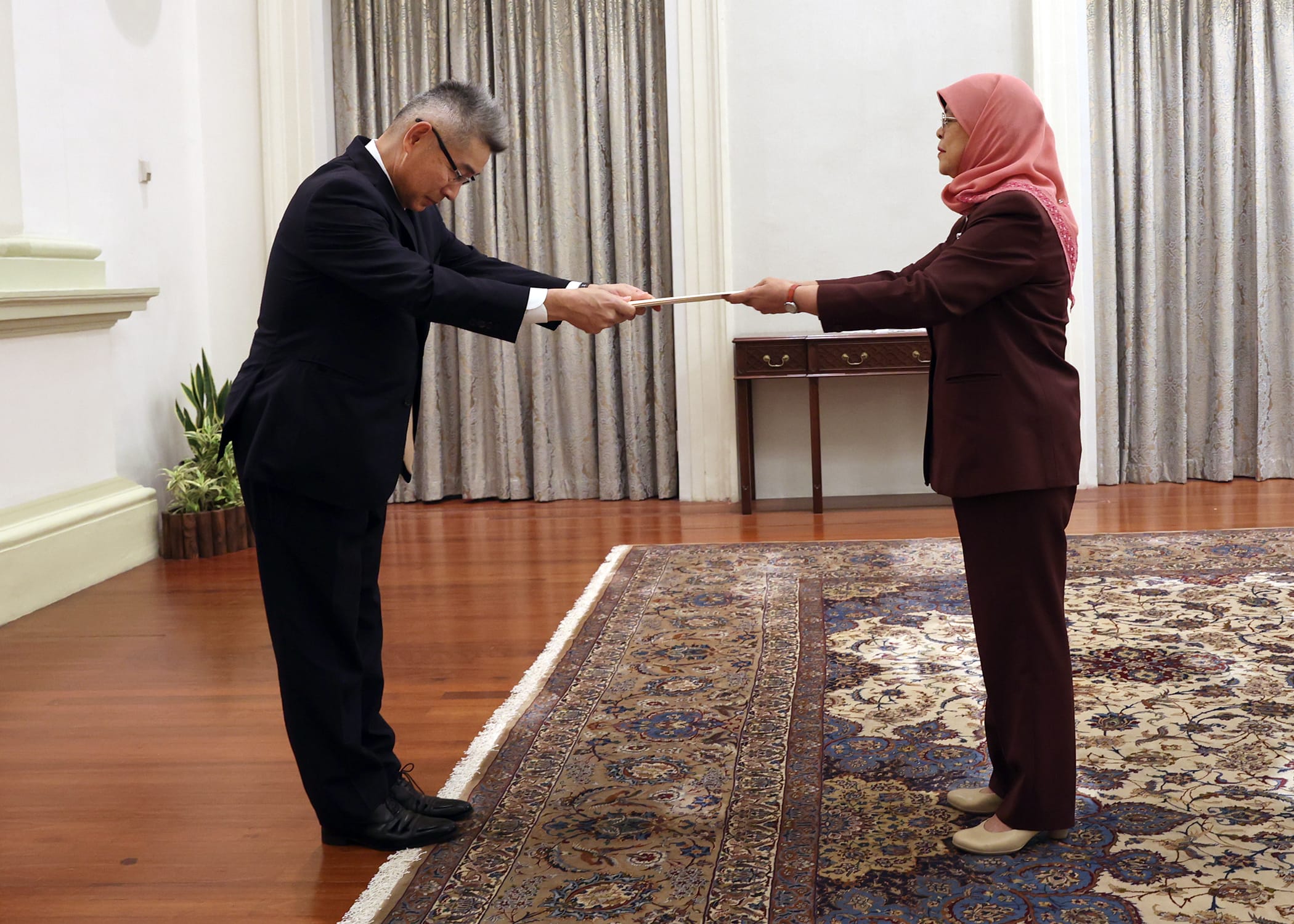 Two people in formal wear exchange document over patterned rug. One wears a pink headscarf.