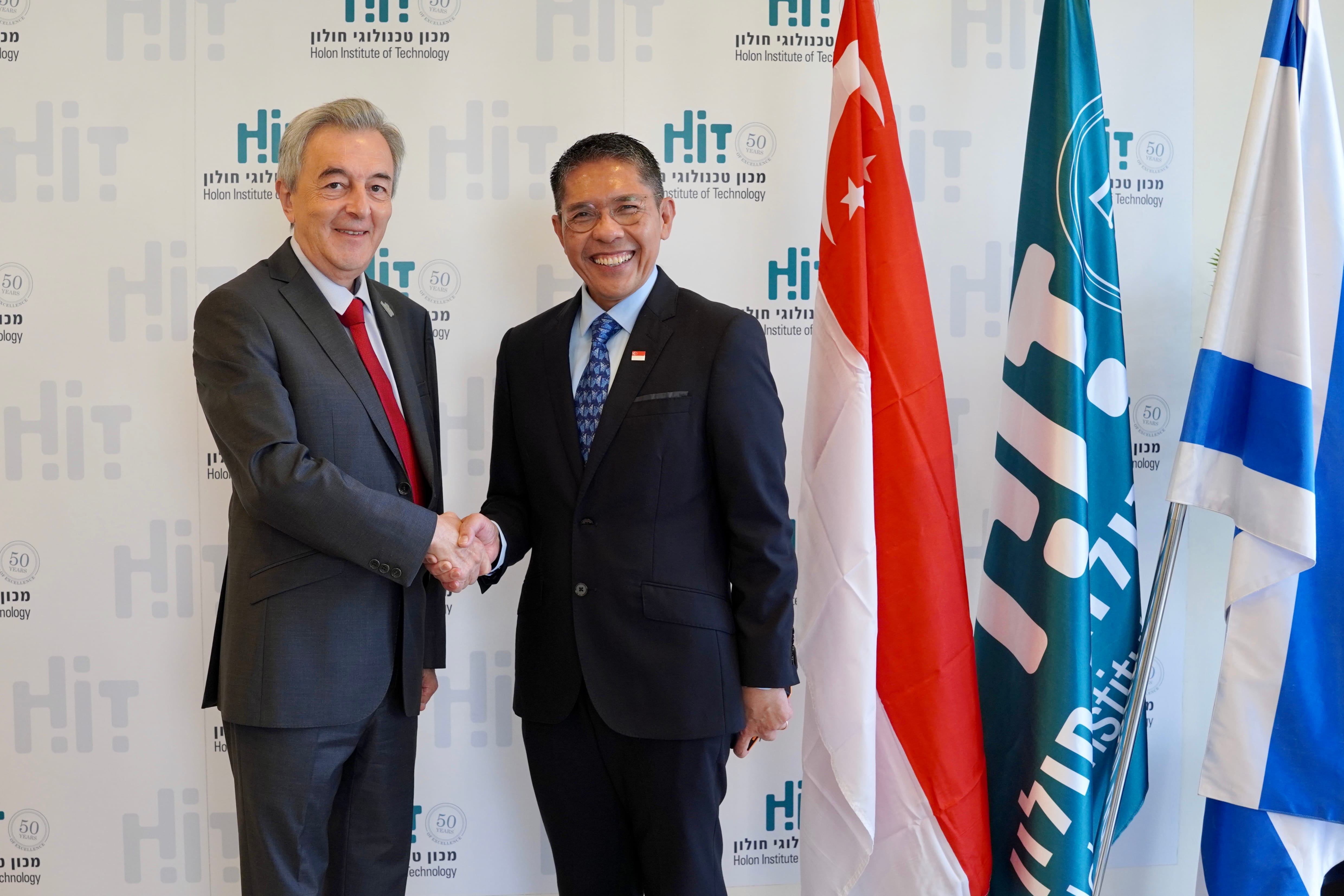 Two men in suits shake hands in front of the Singaporean, HiT, and Israeli flags.