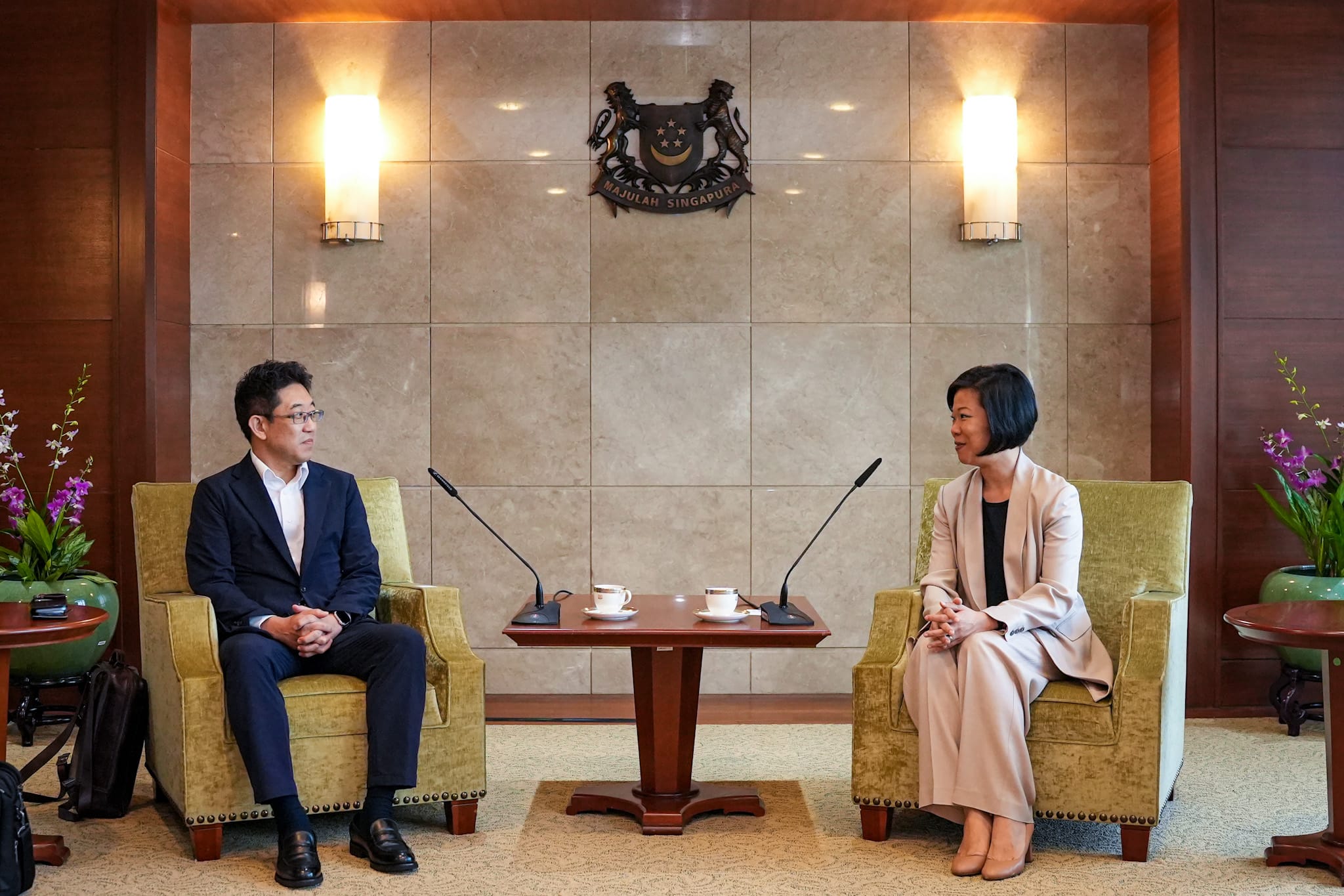 Two people seated facing each other, microphones and tea cups on a table between them. Singapore coat of arms above.