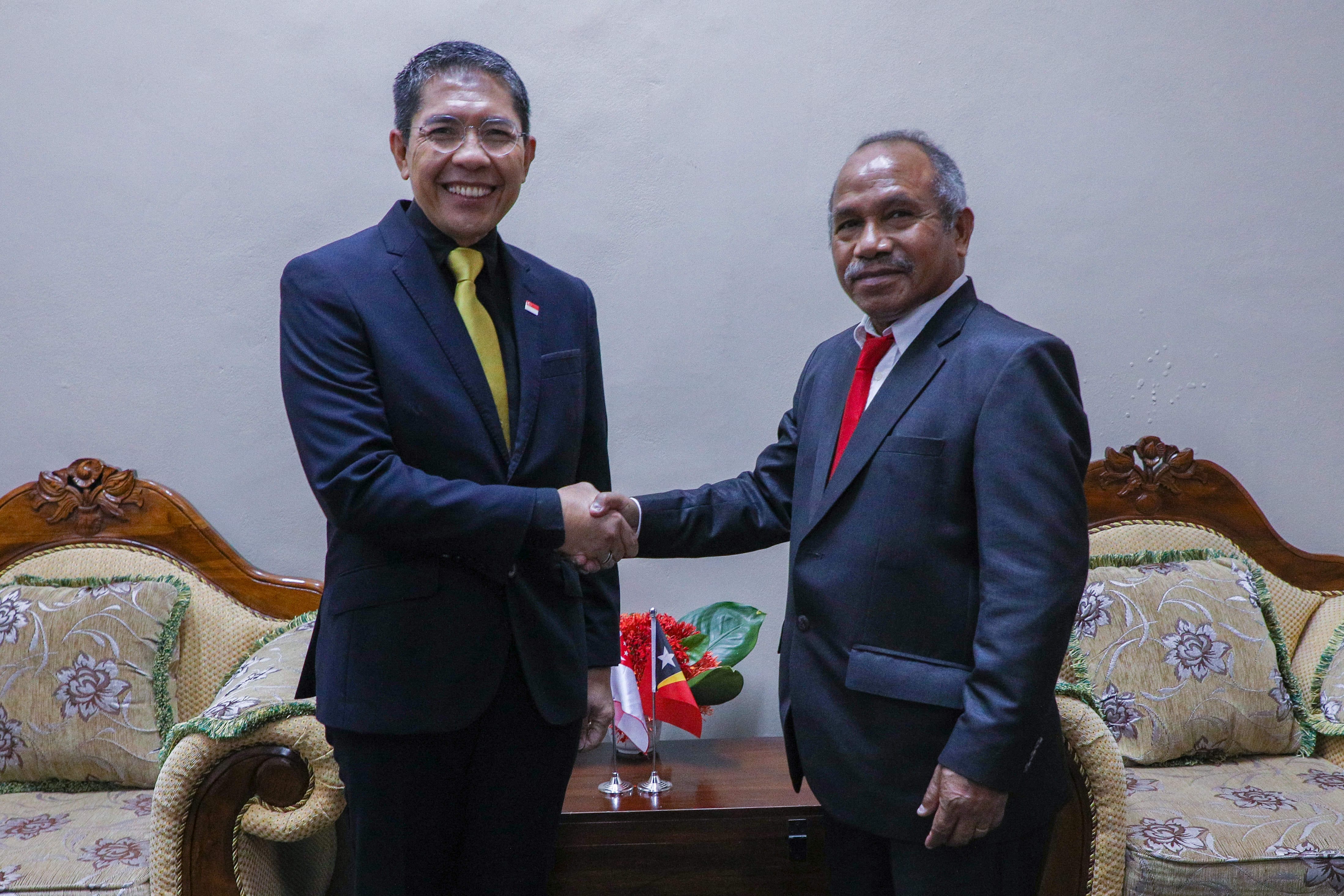 Two men in suits shake hands, a Timor-Leste flag between them.