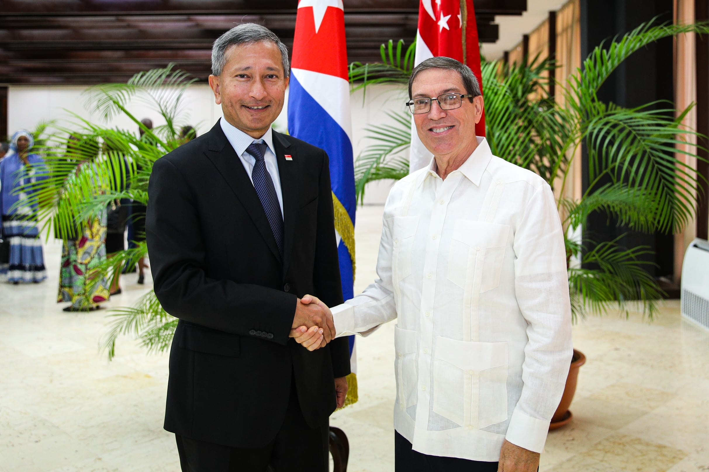 Two men shake hands in front of Cuban and Singaporean flags. One wears a suit, one a white shirt.