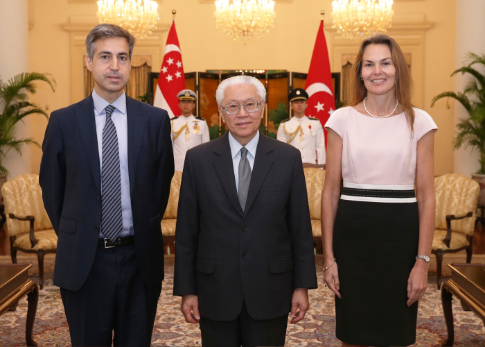 Three people standing in a room with Singapore and Turkey flags in the background.