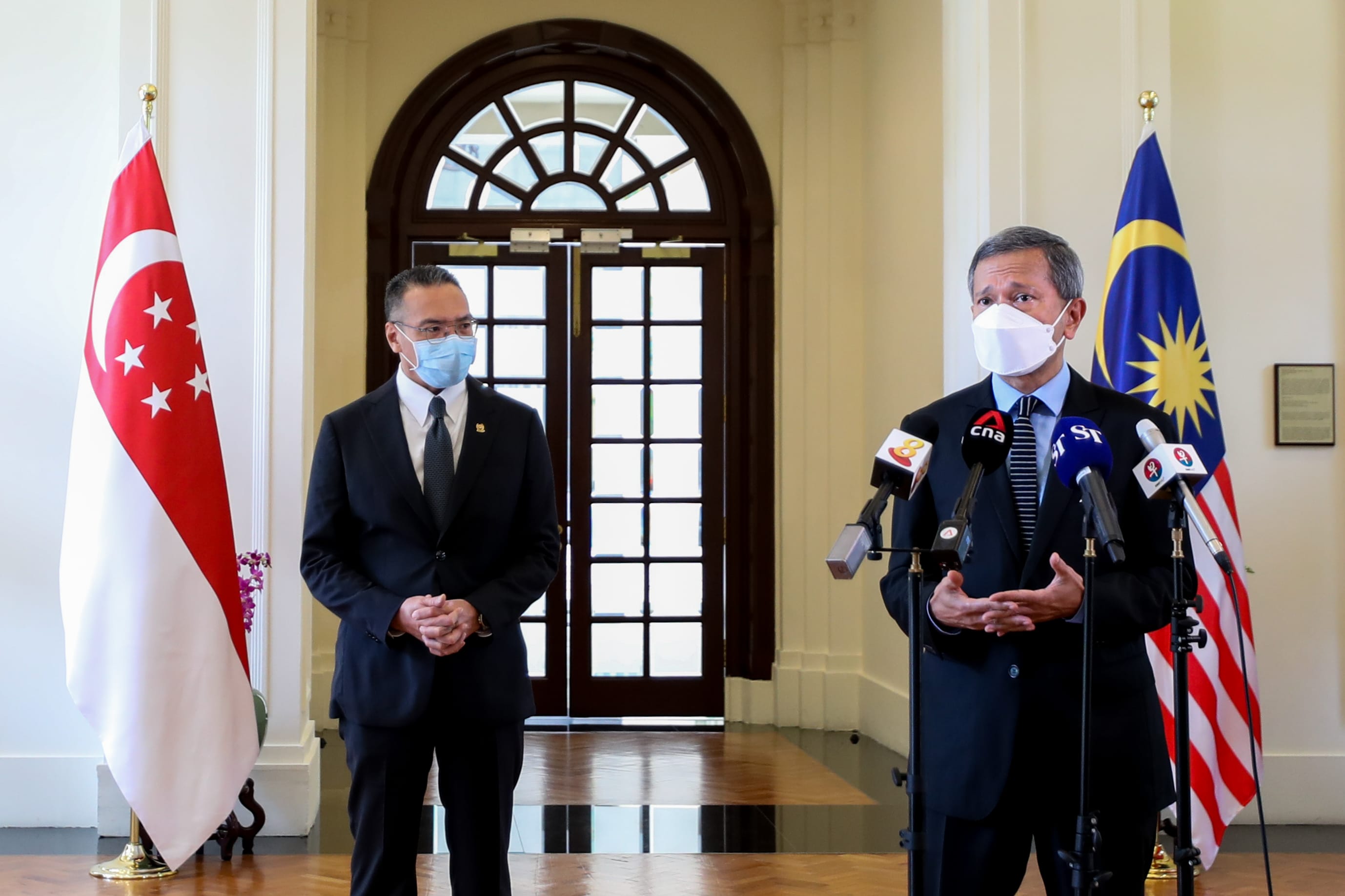 Two men in suits and masks stand before Singapore and Malaysian flags, multiple microphones present.