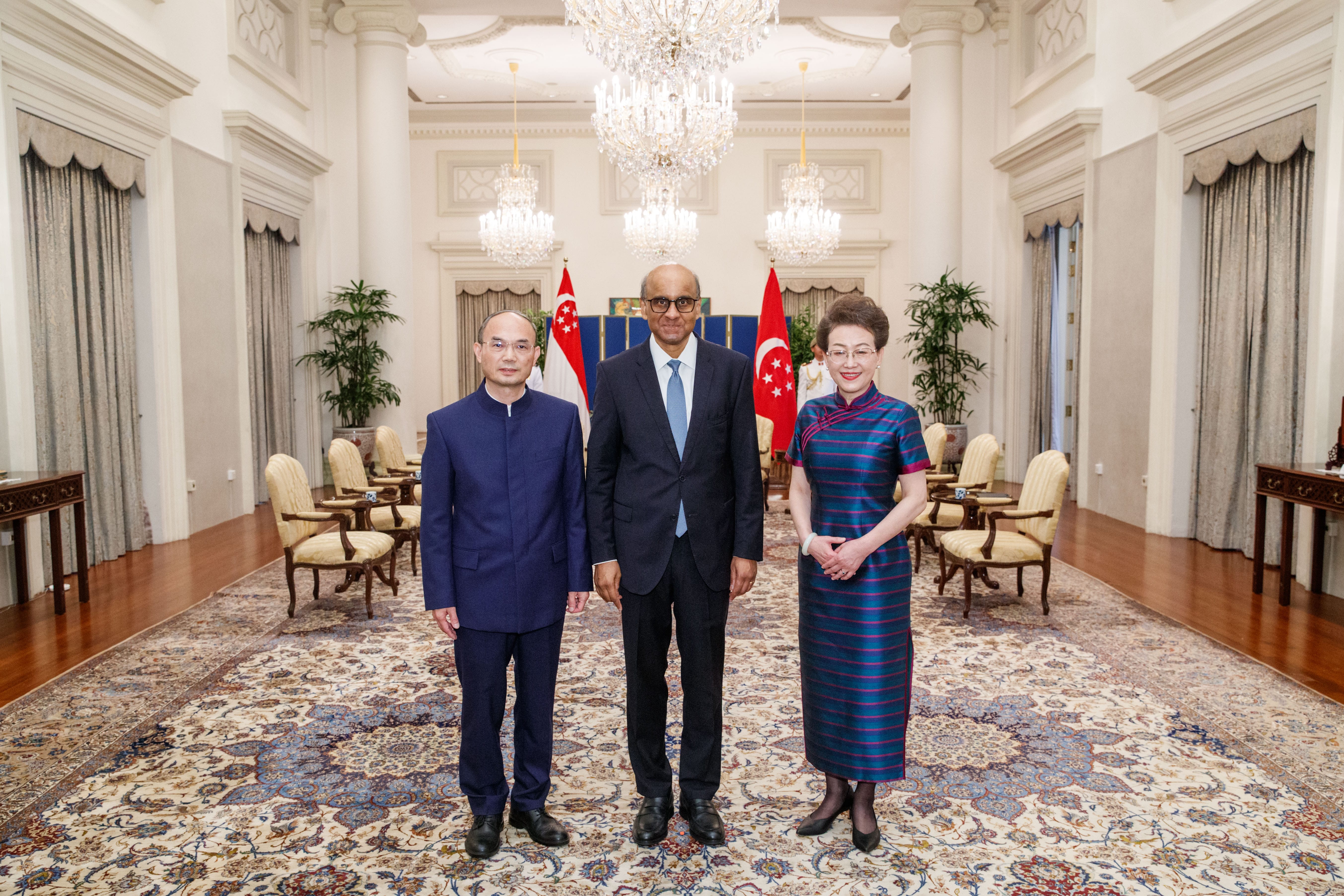 Three people stand in an ornate room with Singapore flags behind them.