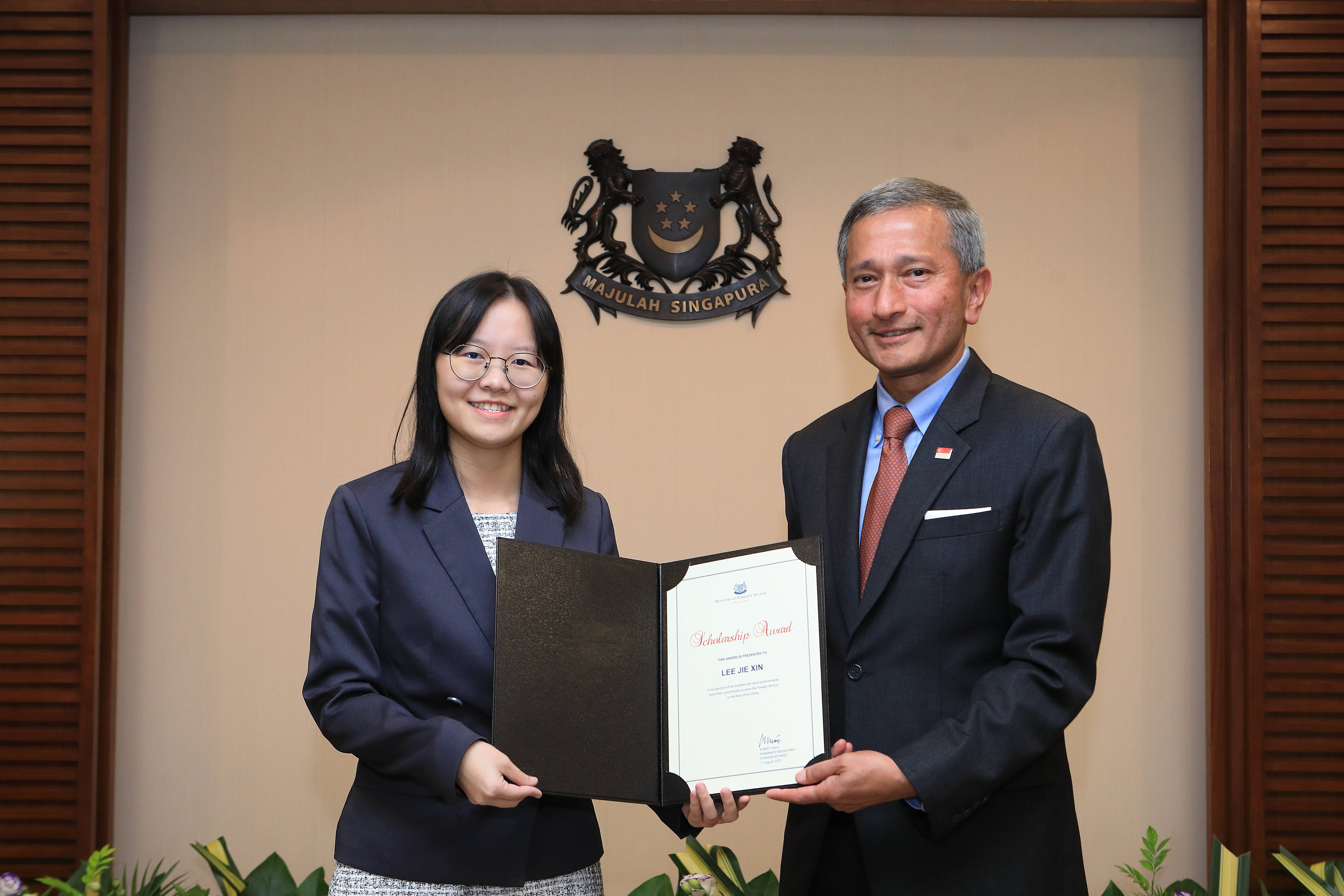 Two people holding a "Scholarship Award" certificate with Singapore's coat of arms in the background.