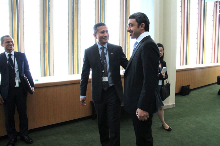 Three people in suits stand in a room with a large window and light green carpet.