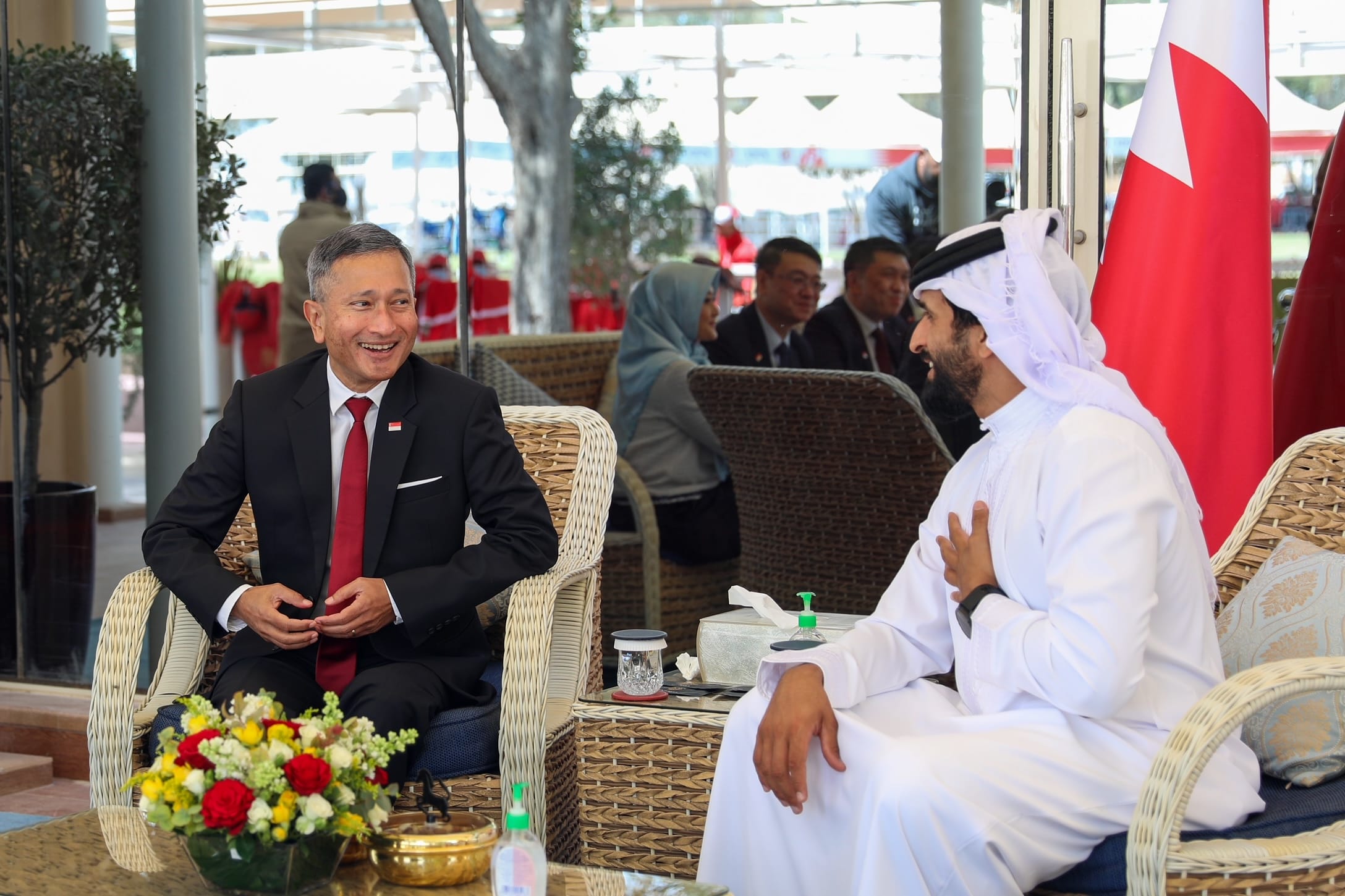 Two men in formal wear sit in wicker chairs, one in a suit and one in thobe, near the Qatar flag.