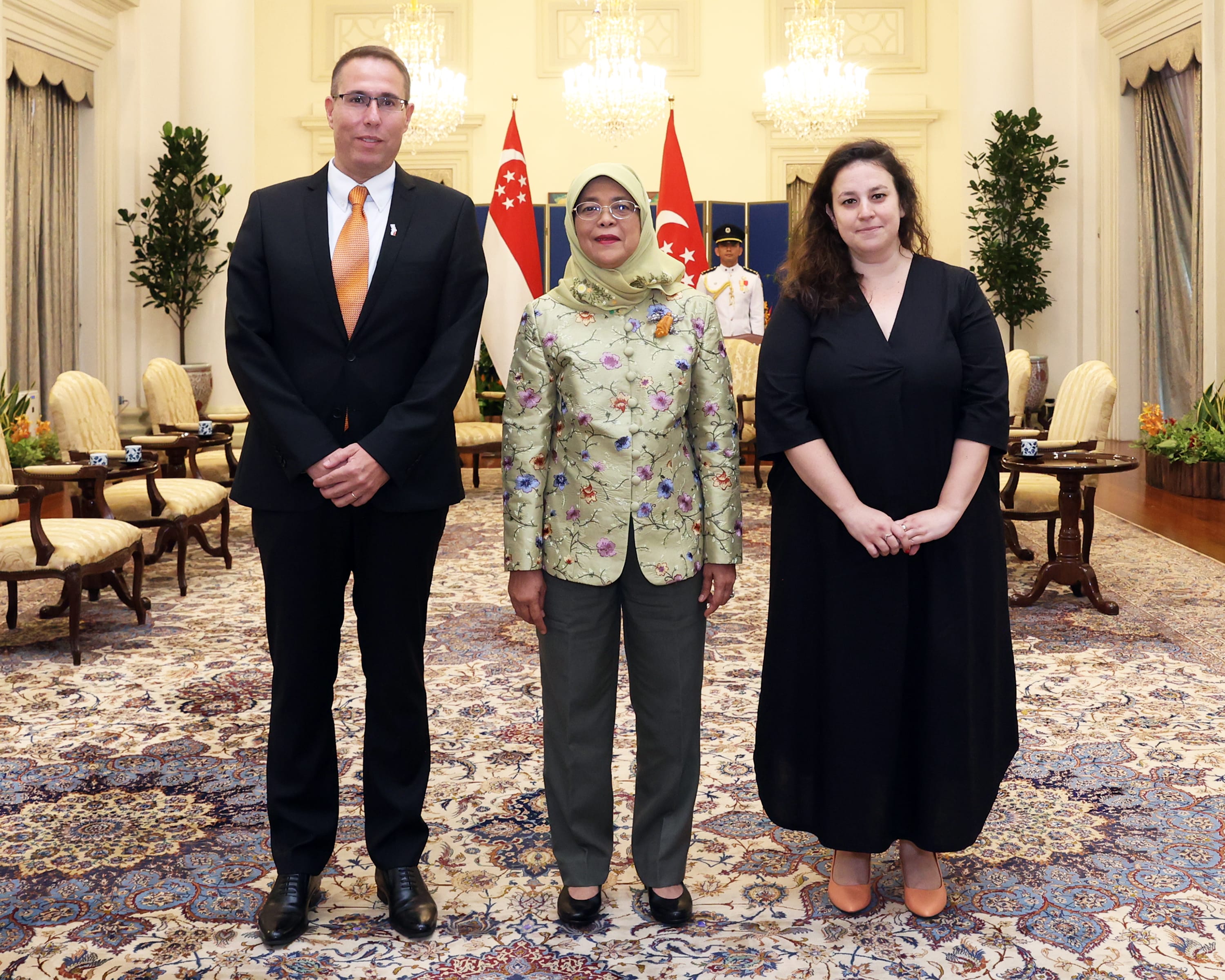 Three people stand in a room with Singapore flags; Halimah Yacob at center.