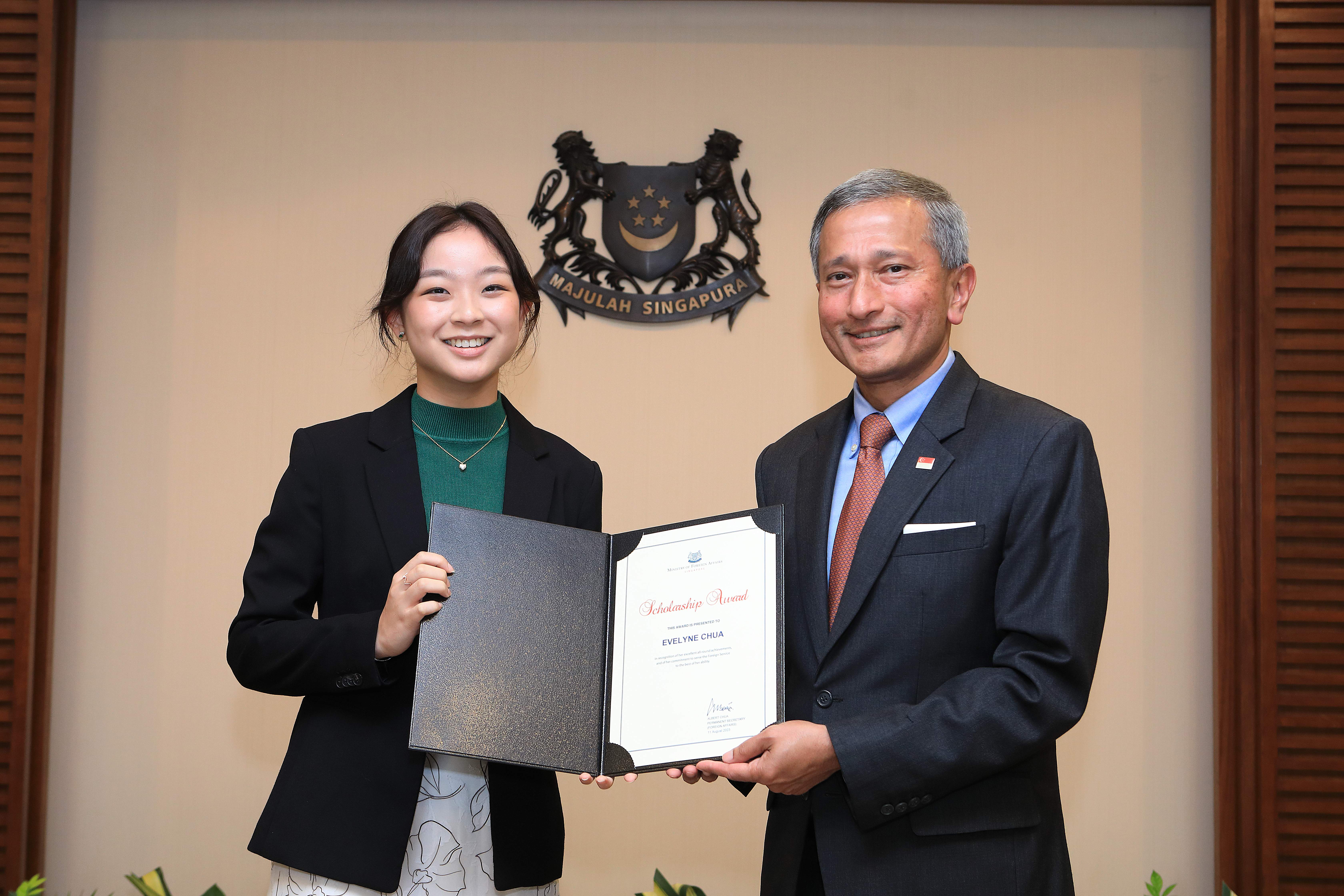 Woman and man in suits holding a scholarship award in front of the Singapore state crest.