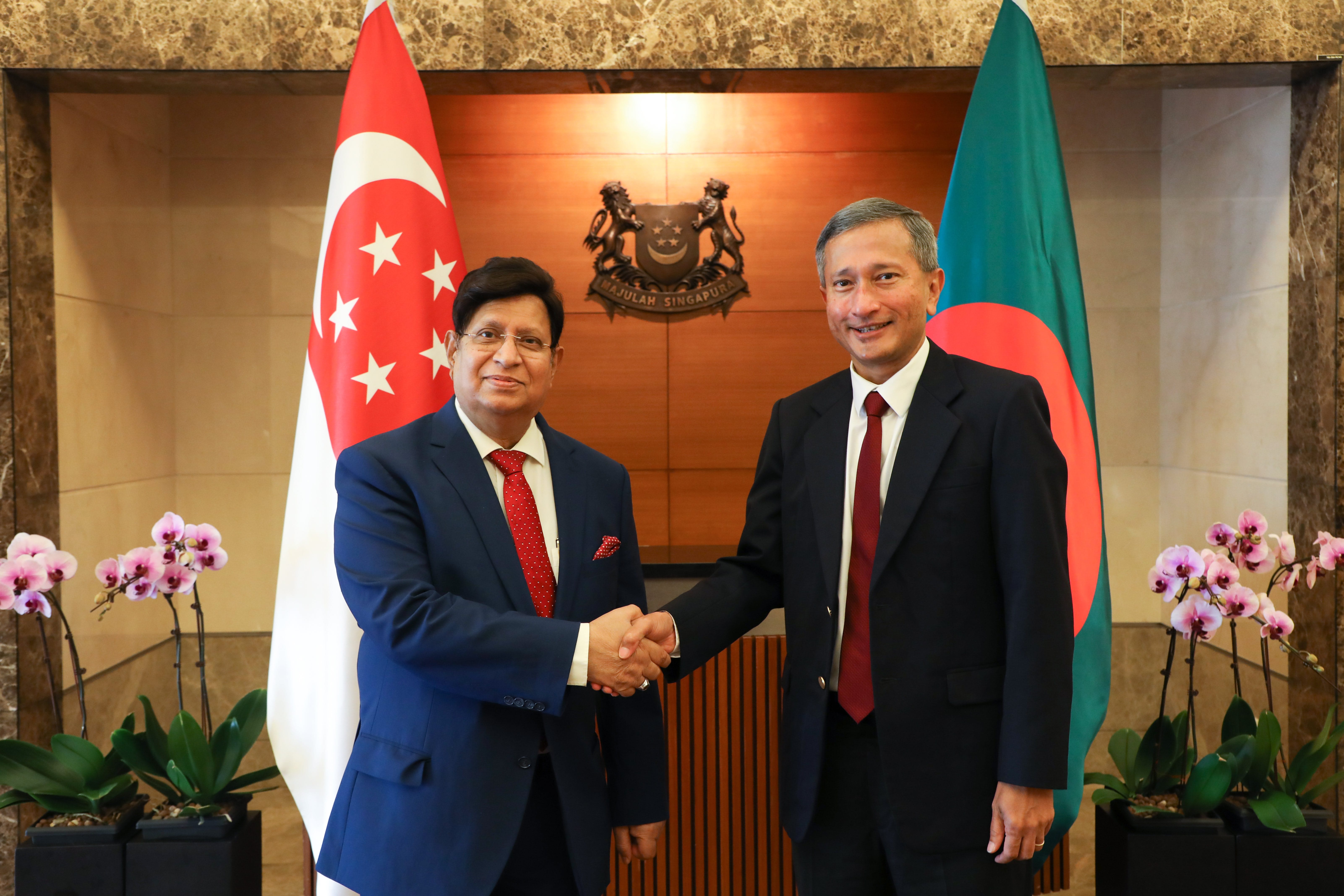 Two men in suits shaking hands, with the flags of Singapore and Bangladesh behind them.