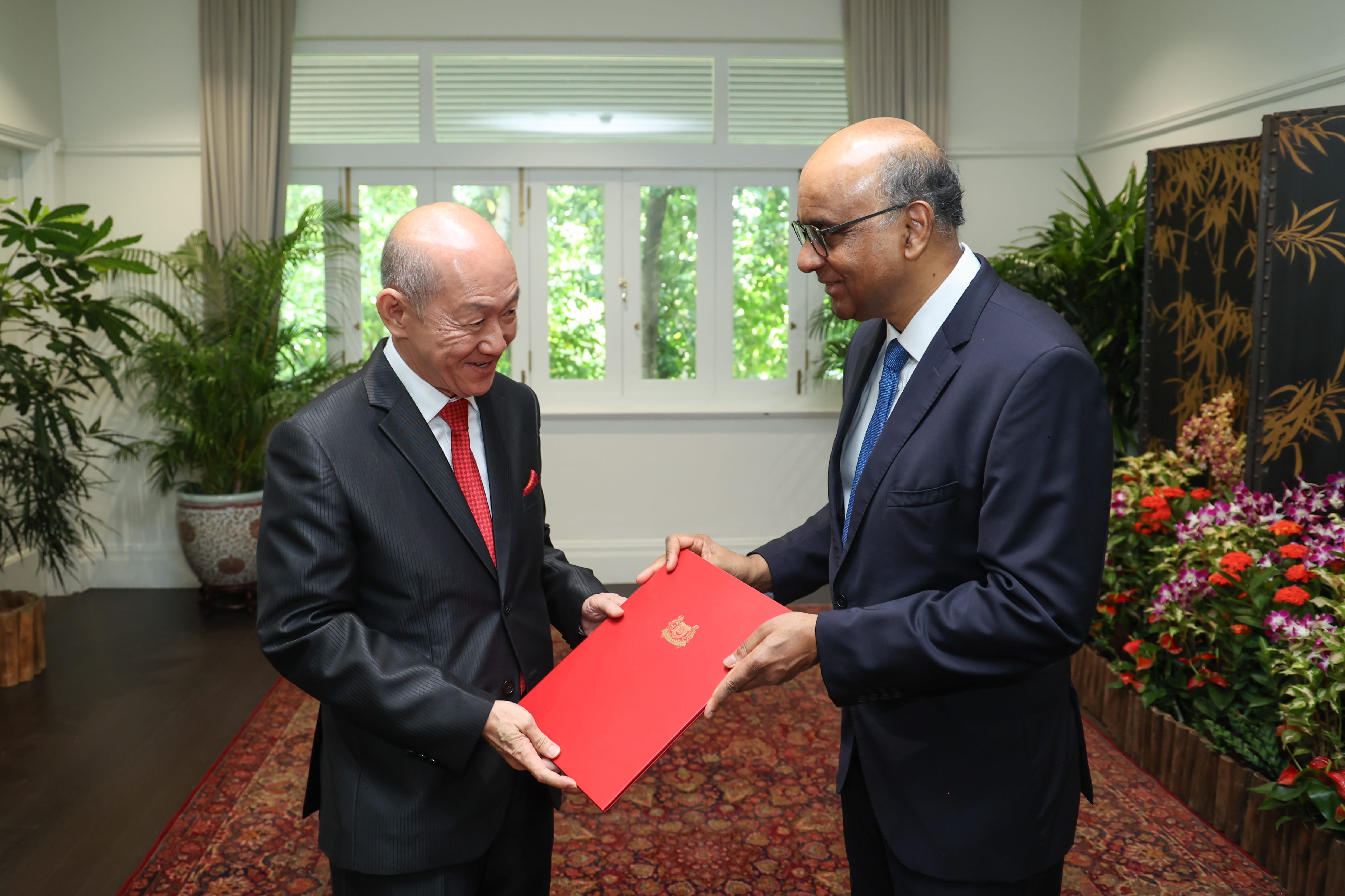 Two men in suits exchange a red folder bearing a seal.