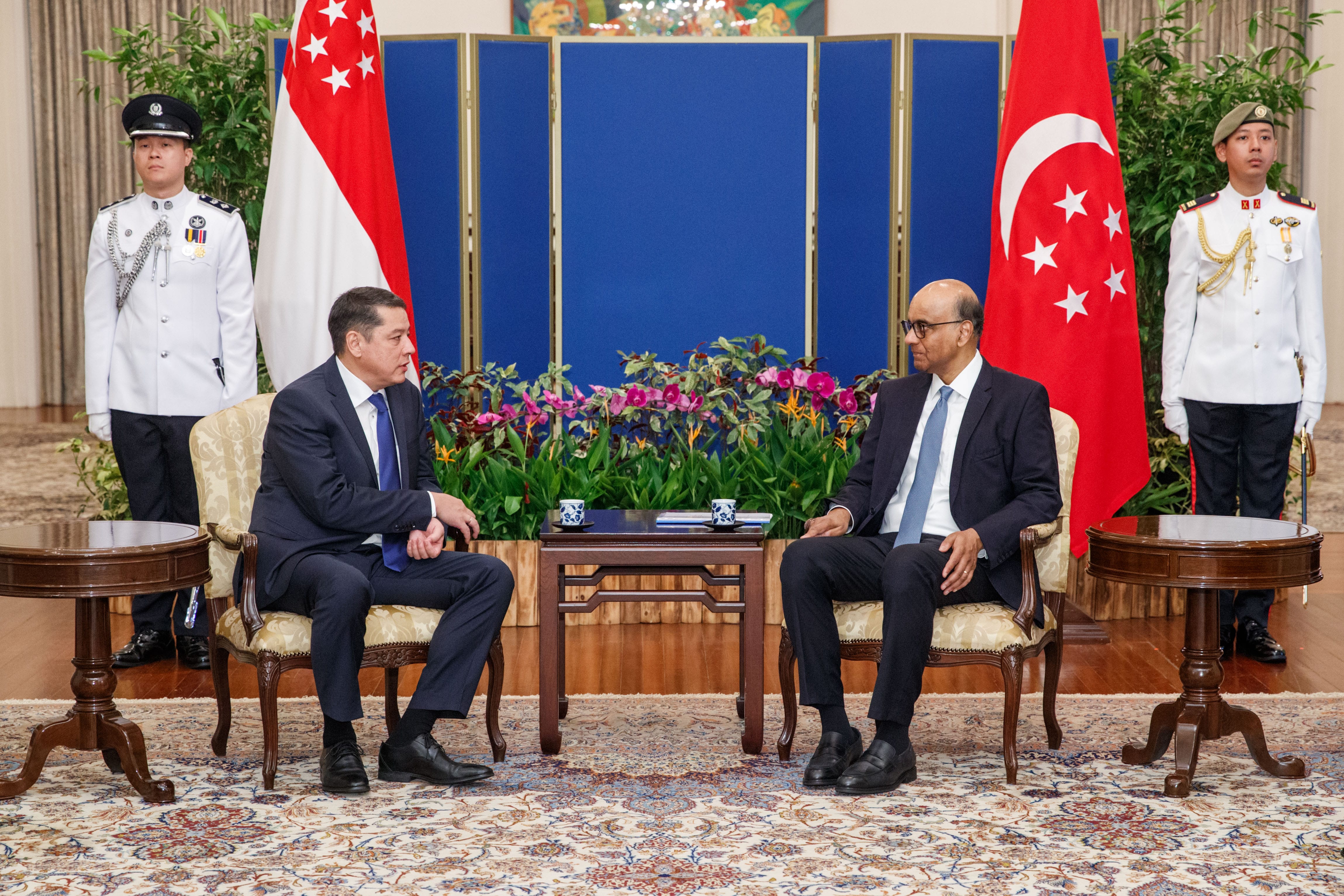 Two men in suits sit opposite each other, flanked by Singapore flags and guards in white uniforms.