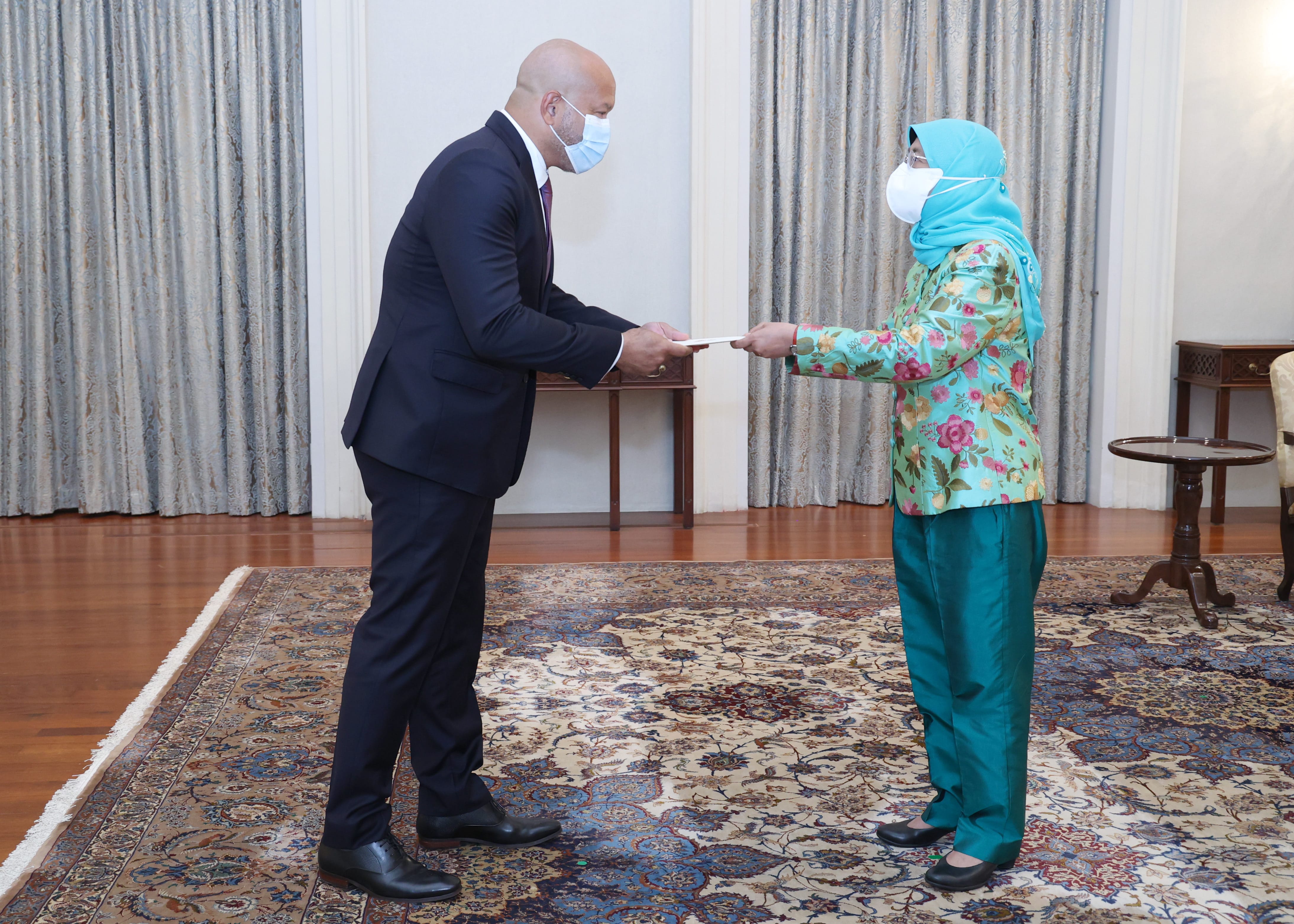 Man in suit and woman in green outfit, both masked, exchanging document.