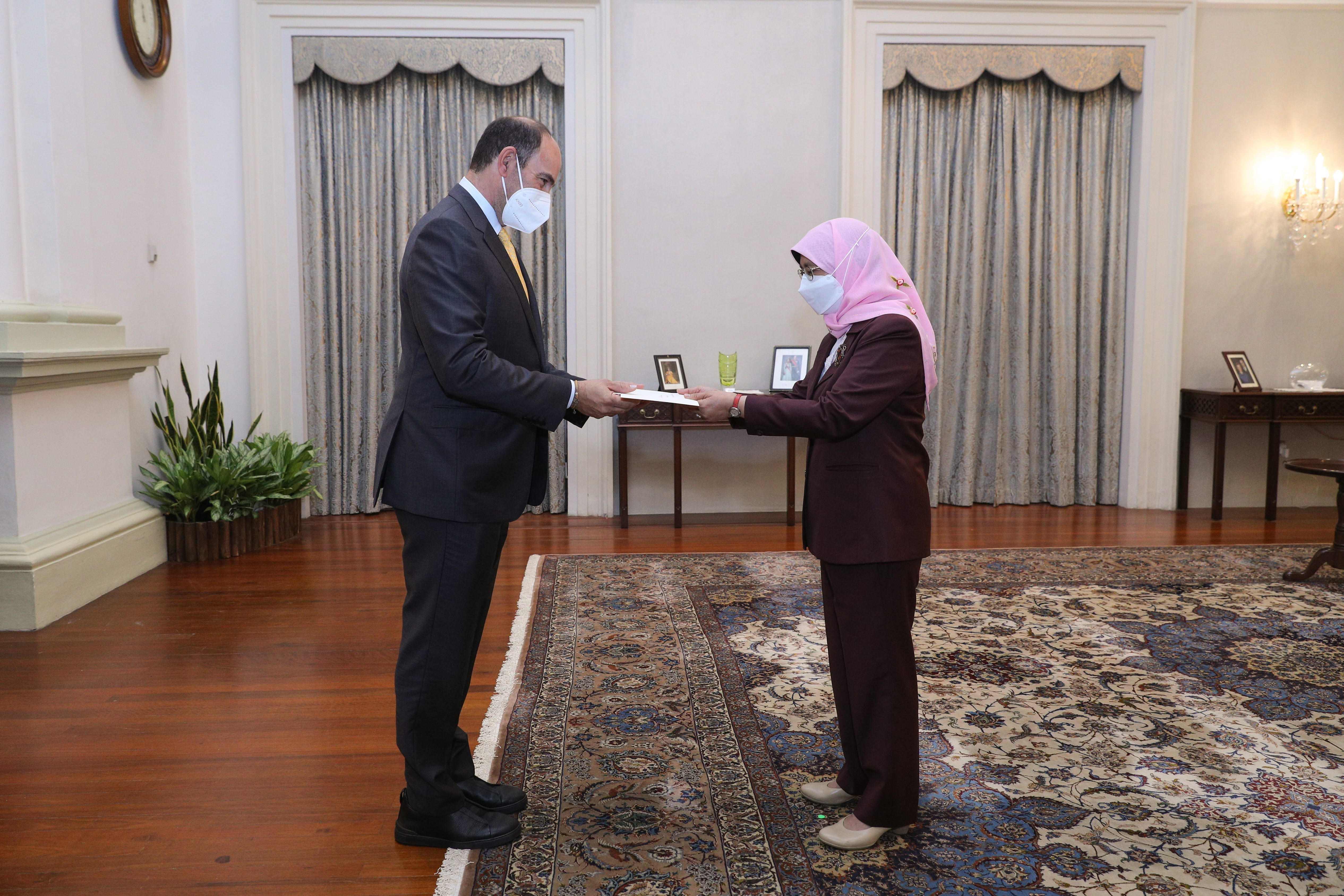 Two people wearing face masks exchange a document in a room with curtains and a patterned rug.