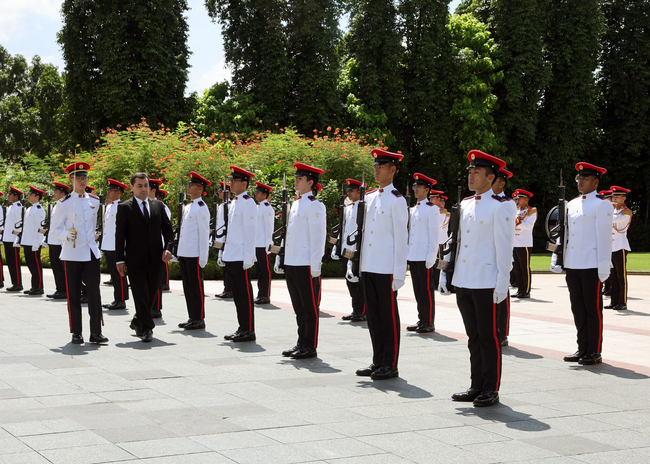 Guard of honor stands at attention in white uniforms and red caps, black-suited official walks by.