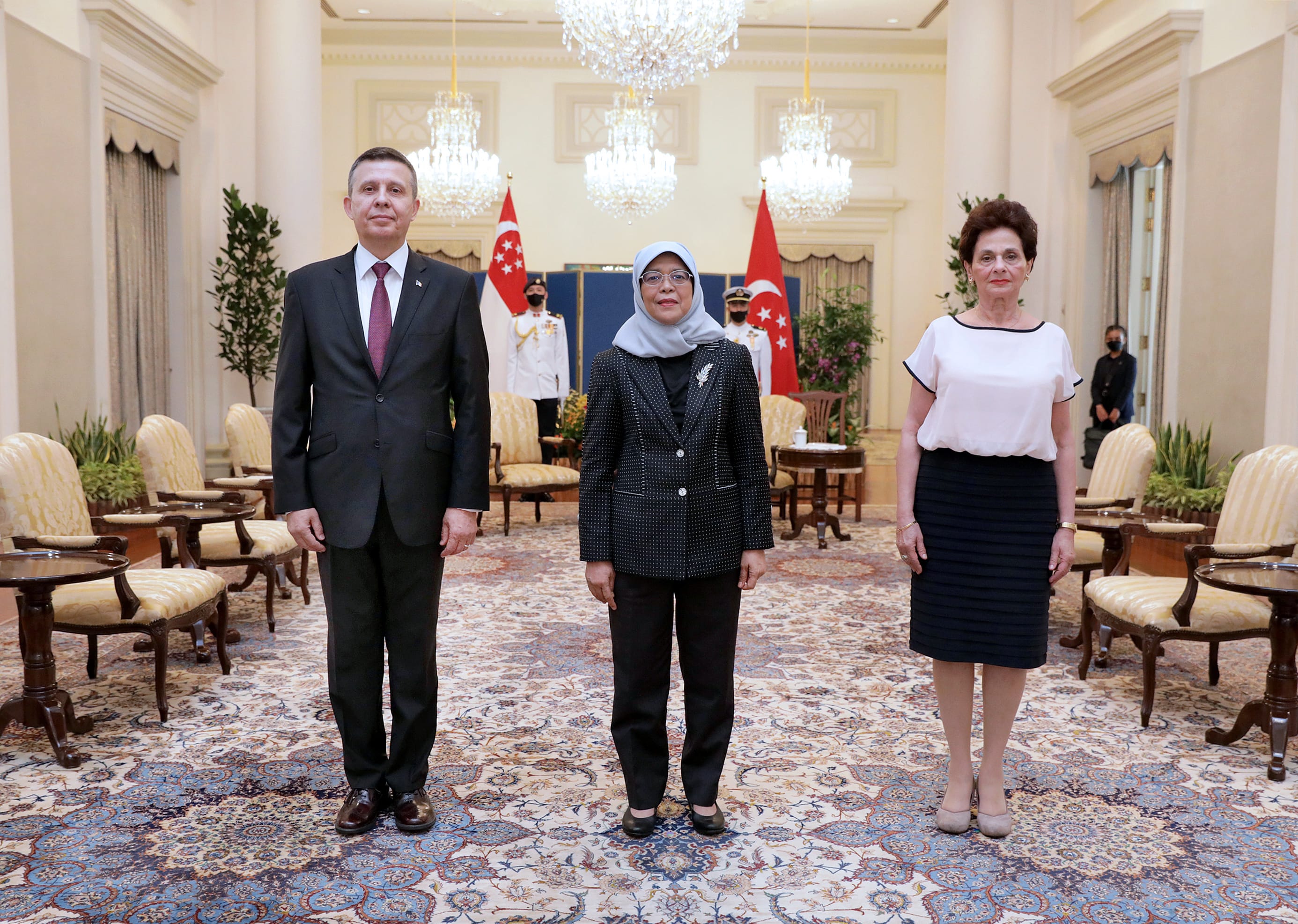 Three people stand in a room with Singaporean flags behind them.
