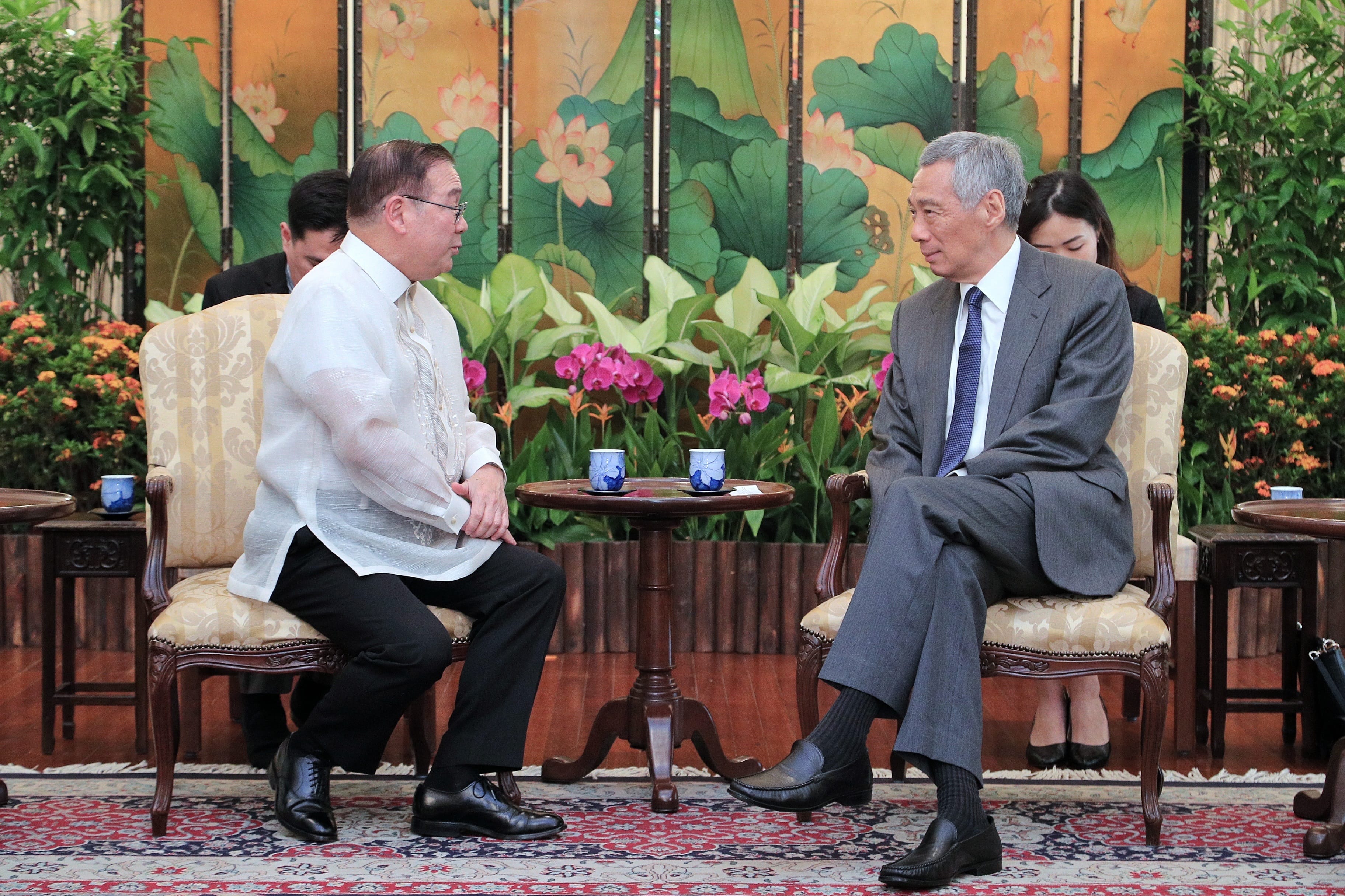 Two men in formalwear sitting in chairs, facing each other, with a floral backdrop.
