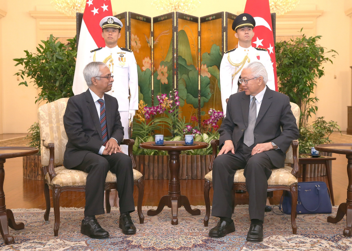 Two men in suits seated, facing each other, flanked by guards and Singapore flags.