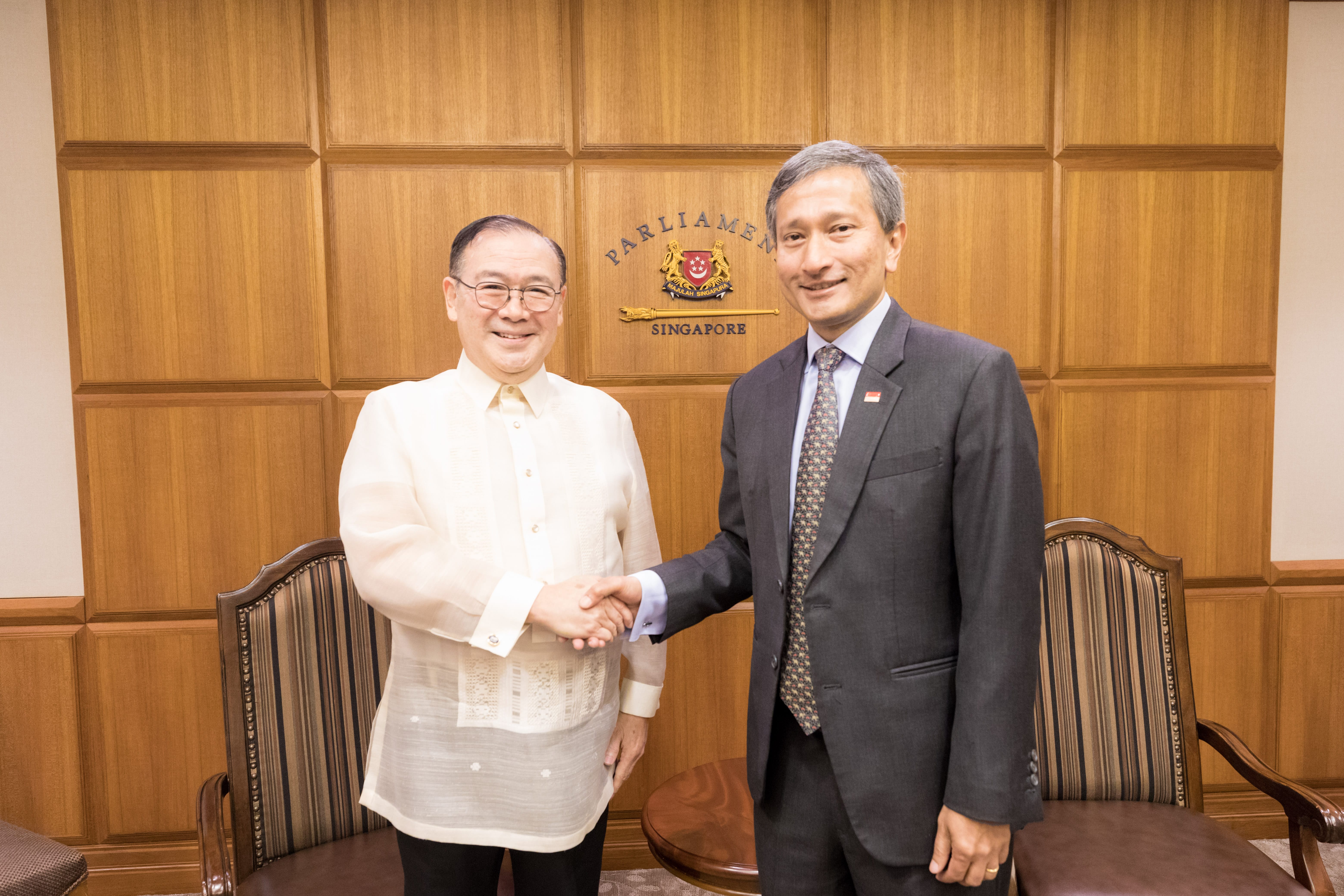 Two men shake hands in front of a wood-paneled wall with the Parliament of Singapore logo.