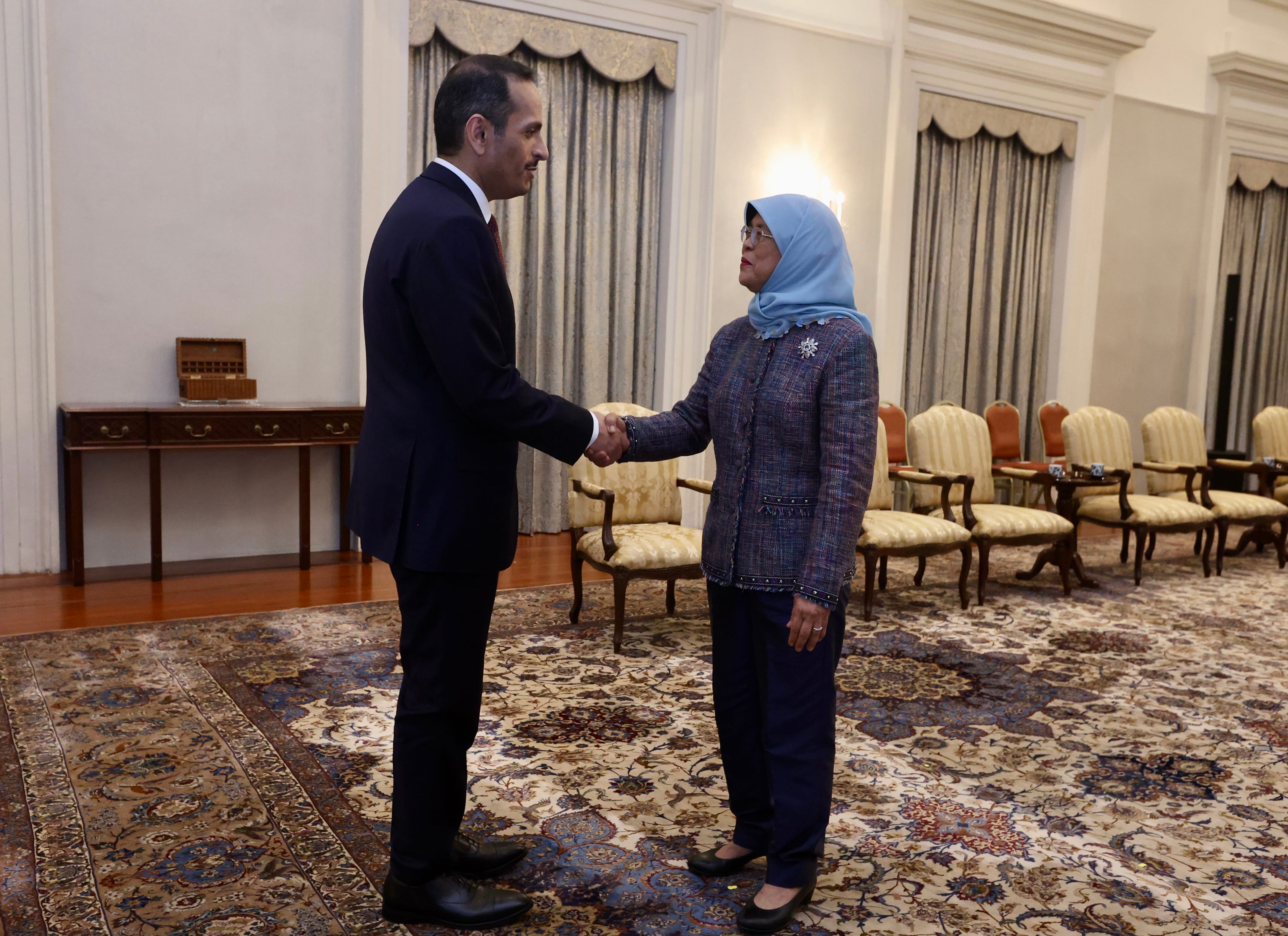 Two people in formal attire shaking hands on an ornate rug in a room with chairs.