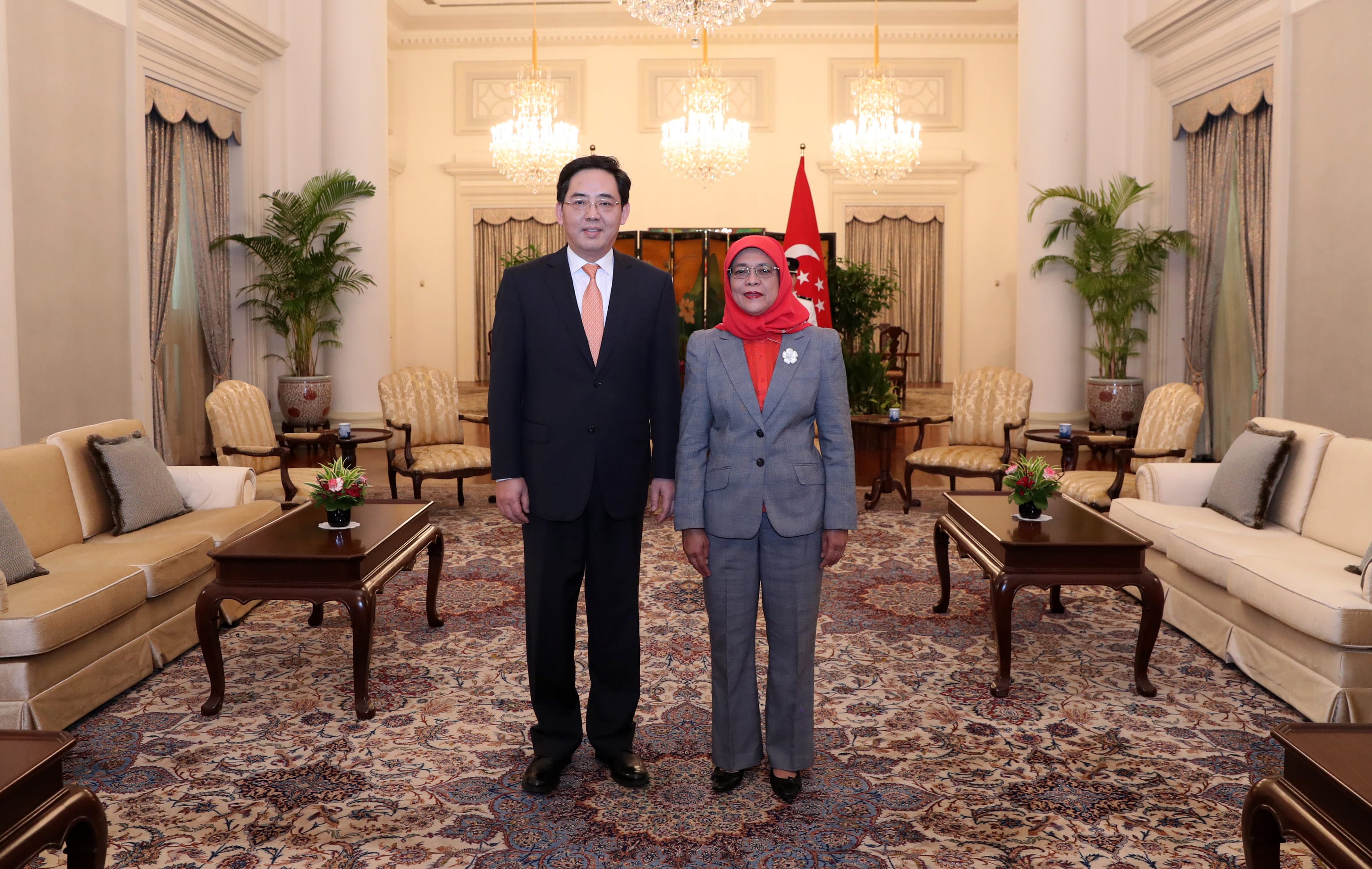 Man in suit and woman in headscarf posing in ornate room, Singapore flag behind.