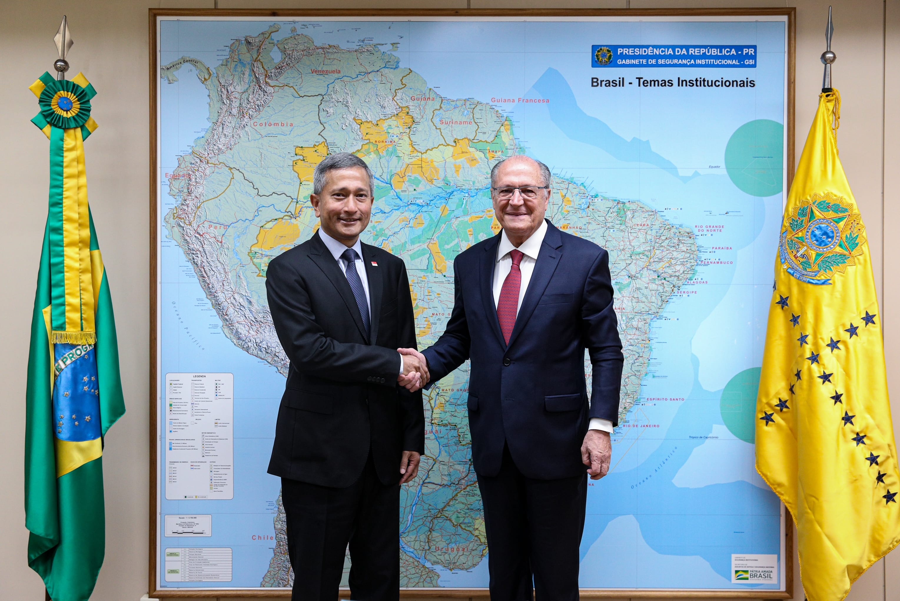 Two men in suits shake hands in front of a map of South America, flanked by Brazilian flags.