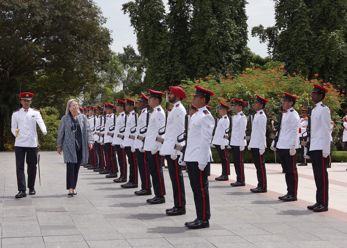 Woman inspecting a long line of uniformed soldiers in white coats and red caps.