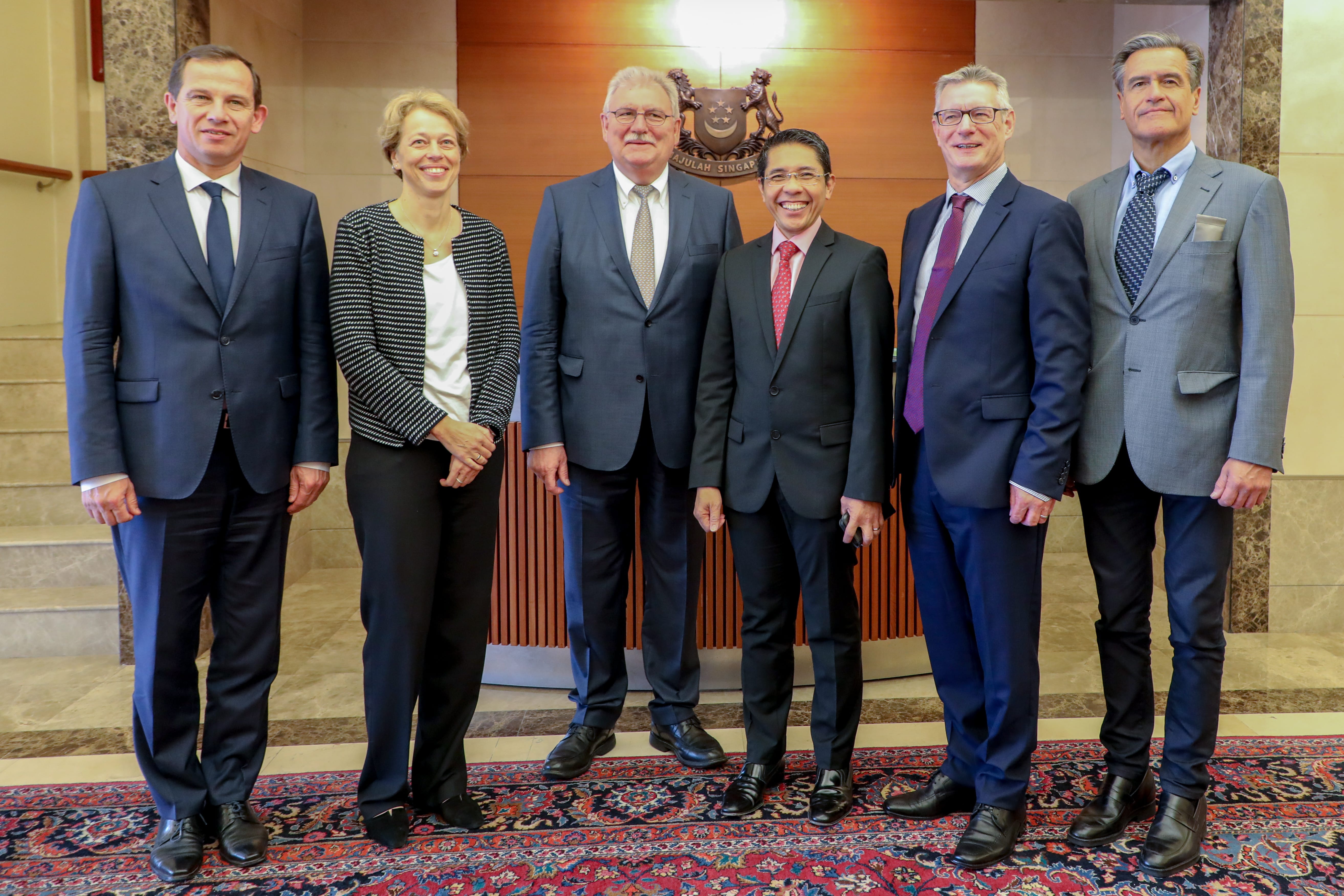 Six people stand formally indoors, in suits, against a wooden backdrop with a coat-of-arms.