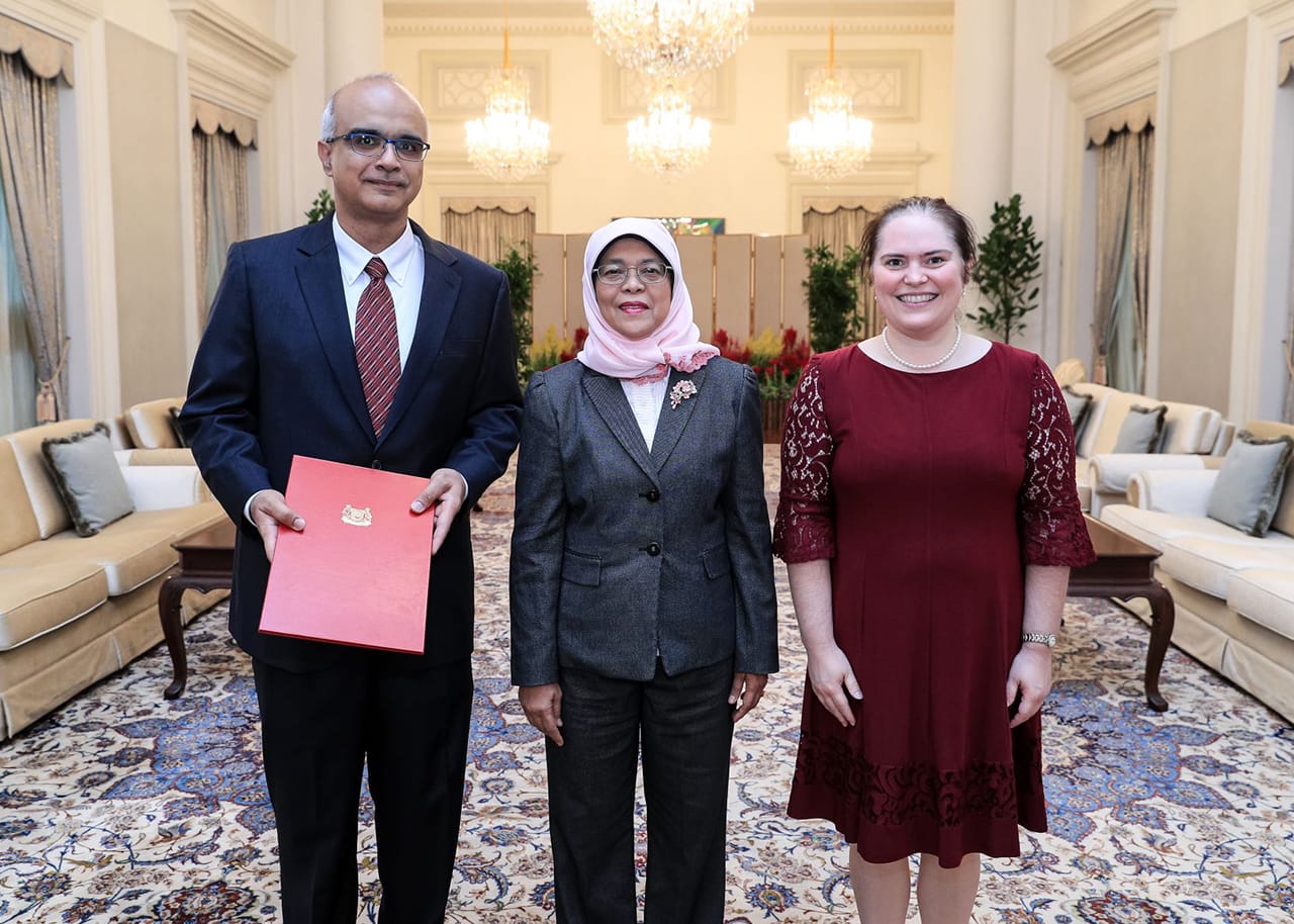 Three people posing indoors; man holds red folder with crest, woman in hijab, woman in dress.