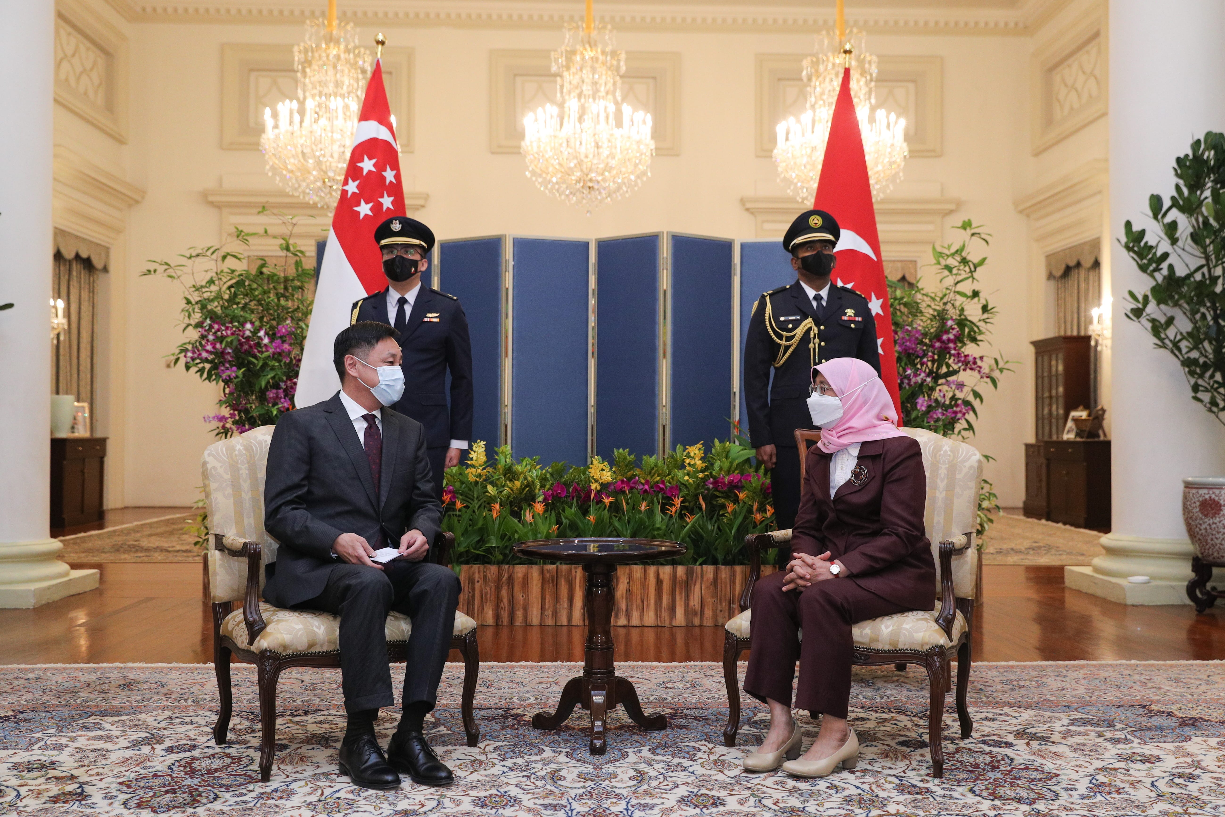 Two people seated, two guards standing near Singapore flags, wearing masks.