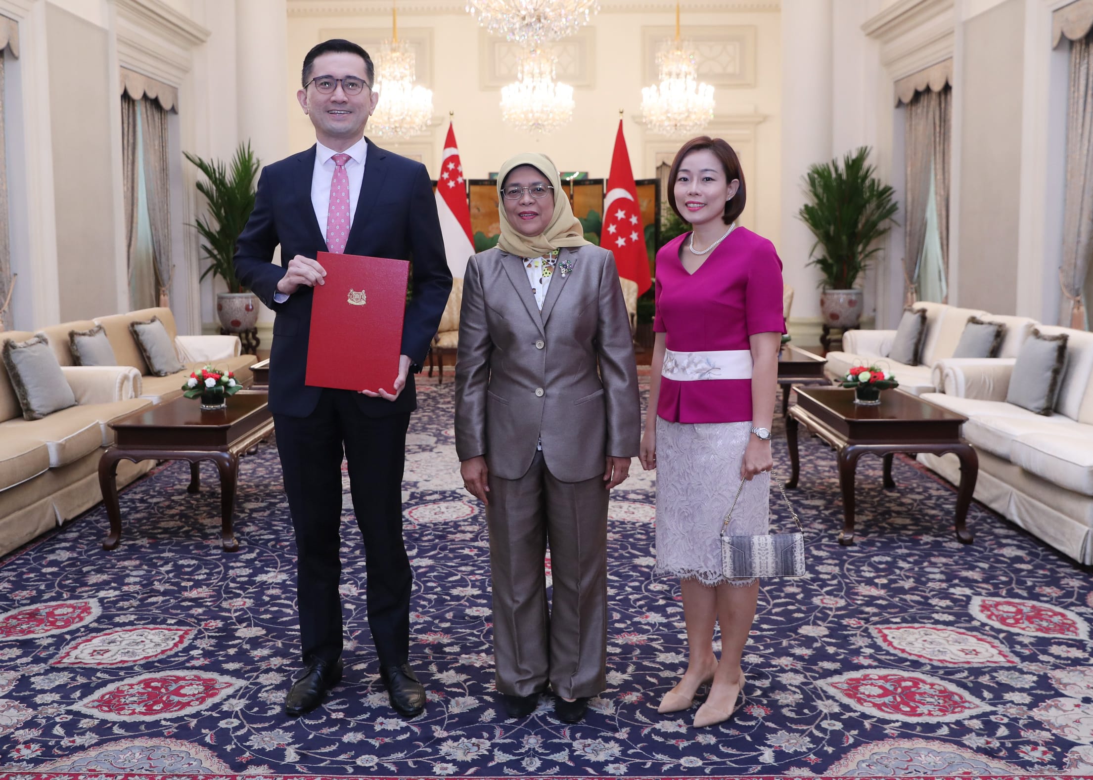 Three people posing indoors with Singapore flags, patterned rug, chandelier. Man holds red document.
